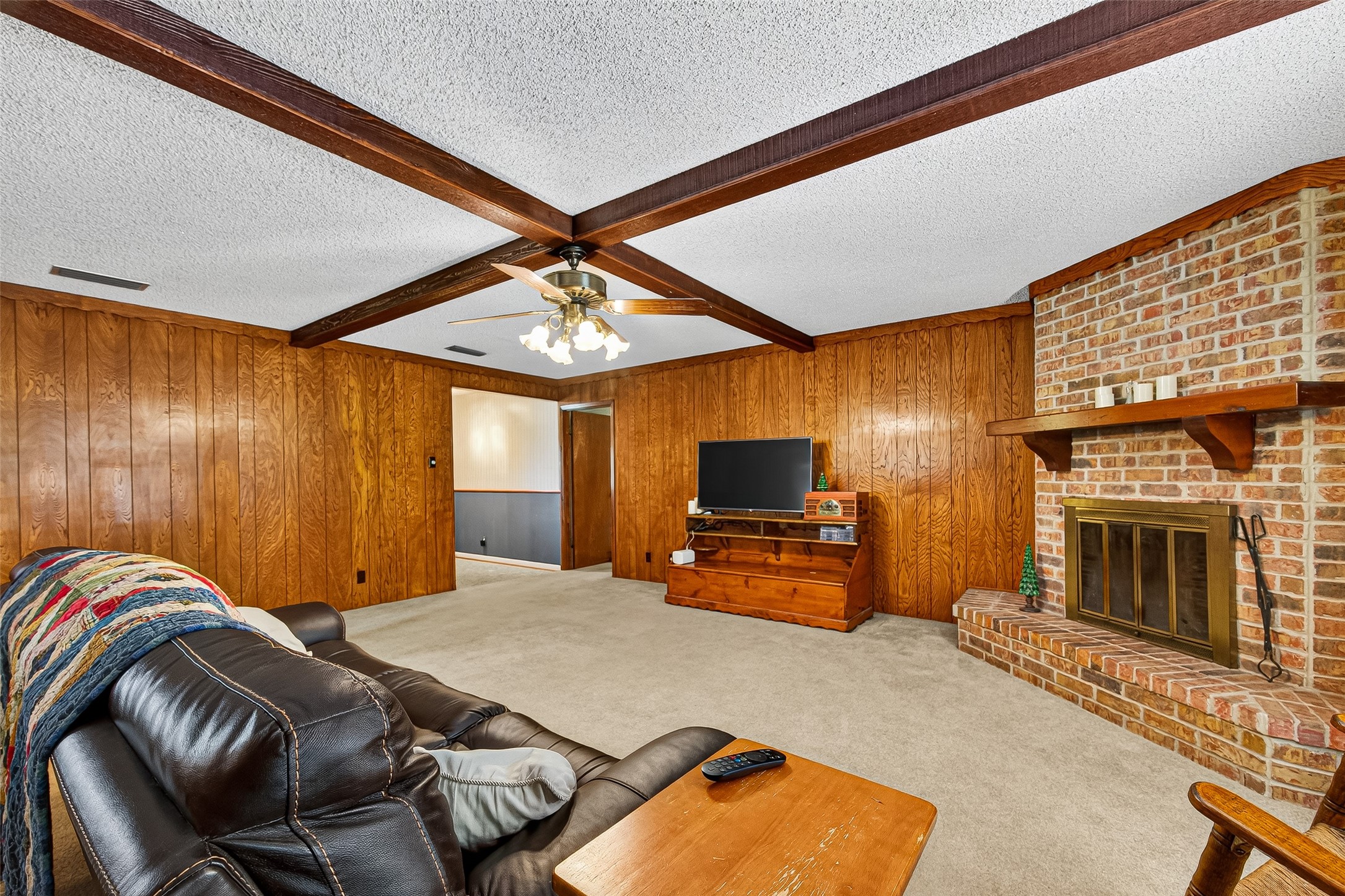This cozy living room features wood-paneled walls and a charming brick fireplace. It has a ceiling fan, exposed beams, and ample space for seating, making it perfect for relaxation and gatherings.
