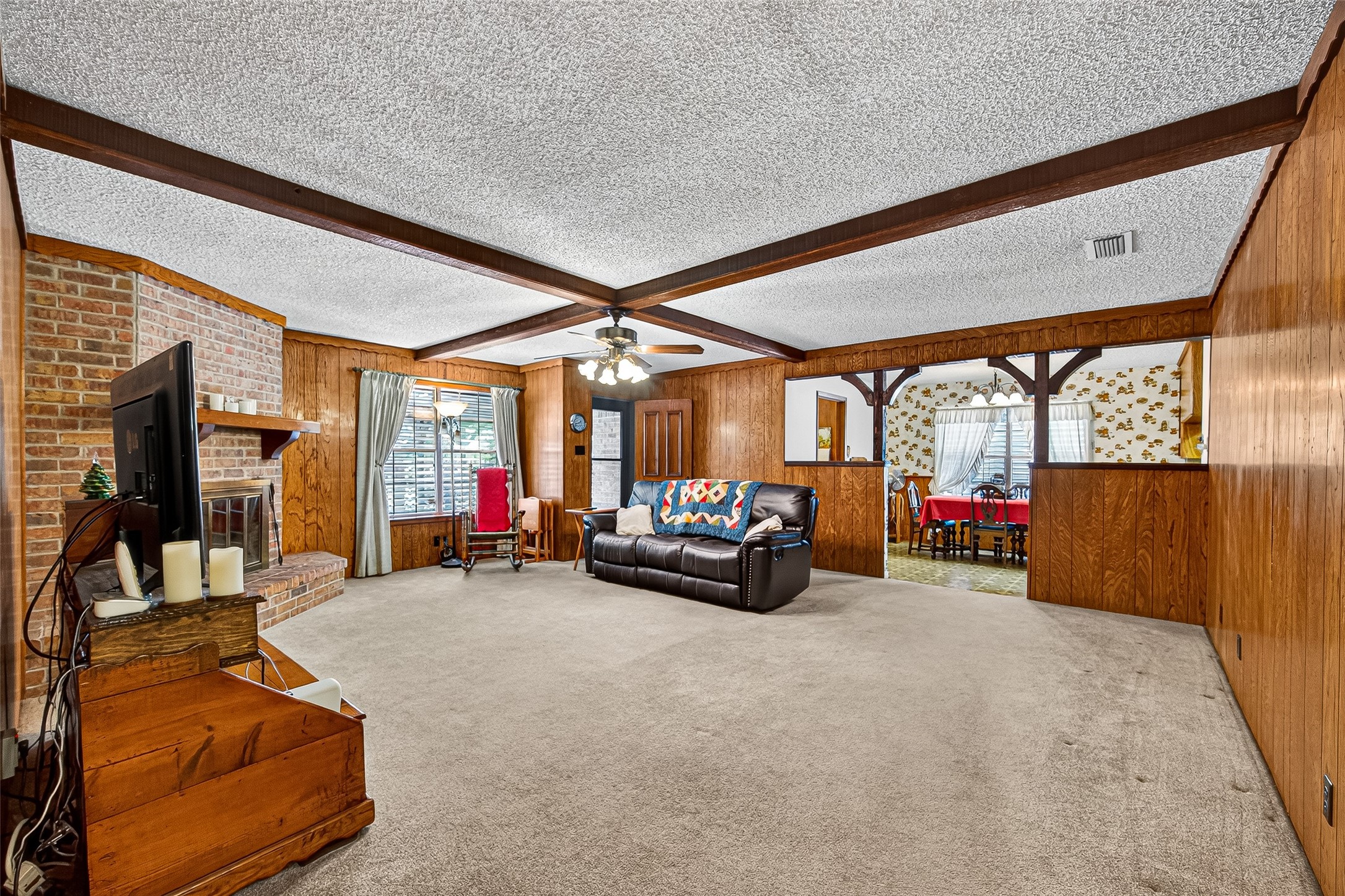 This cozy living room features wood-paneled walls, a brick fireplace, and beamed ceiling accents. It opens to a dining area with a large window, offering a warm and inviting space for gatherings.