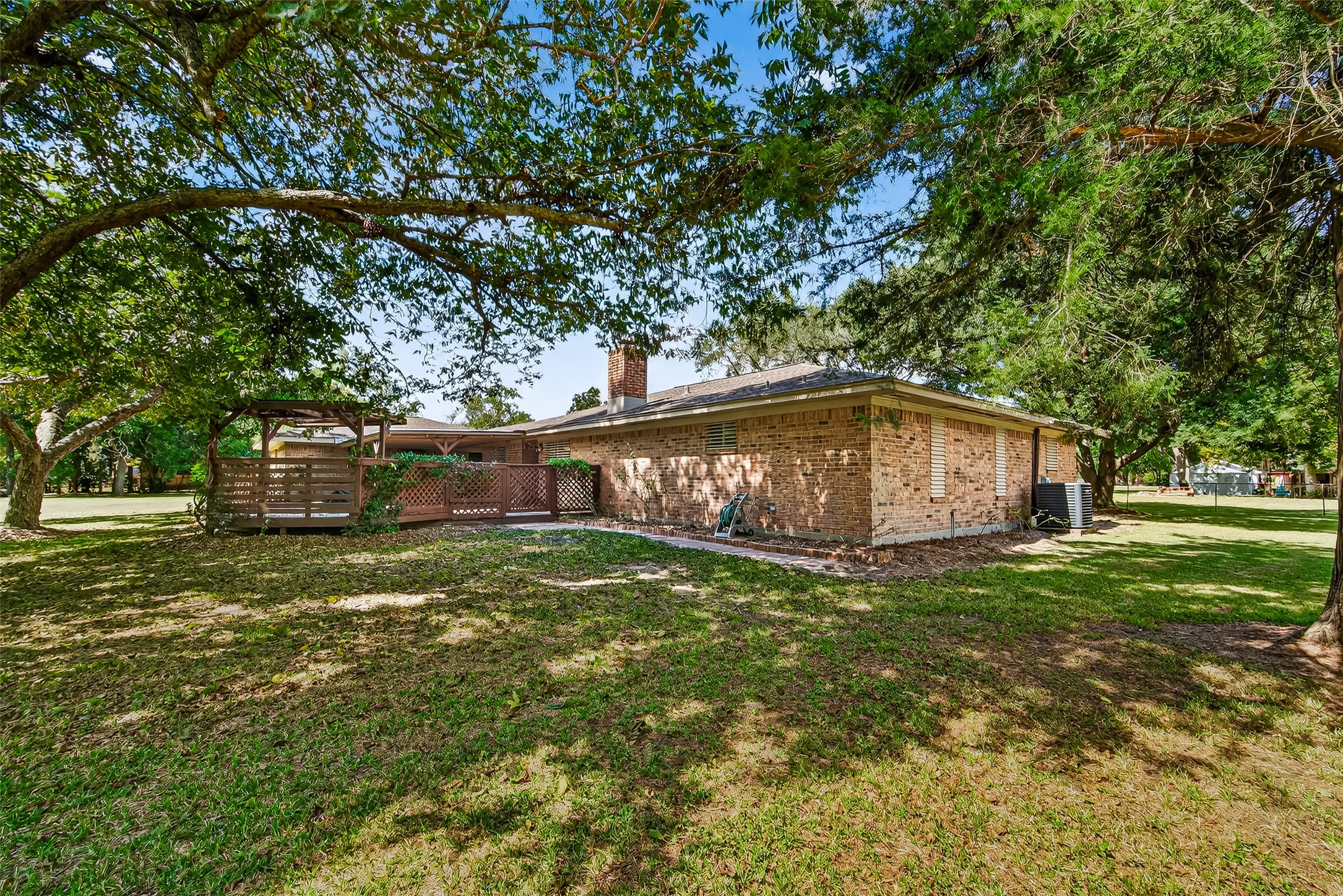 This photo shows the backyard of a brick house with a spacious lawn. There's a wooden deck or pergola attached to the house, offering a cozy outdoor space. Hot tub ready for installation. The setting is peaceful, with plenty of greenery.