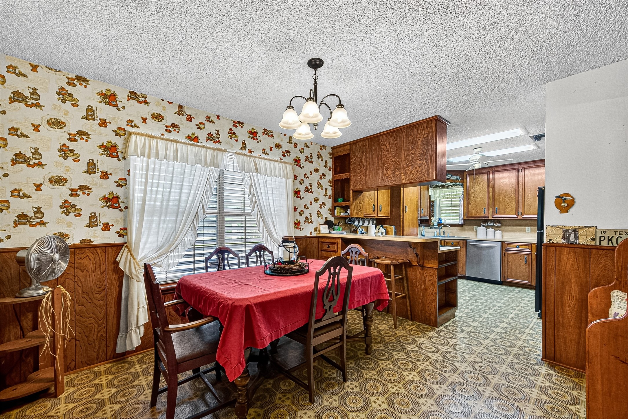 This cozy dining area features vintage floral wallpaper and wood paneling, creating a warm, nostalgic atmosphere. It has a spacious table, abundant natural light from large windows, and an open view into a well-equipped kitchen with wood cabinetry.