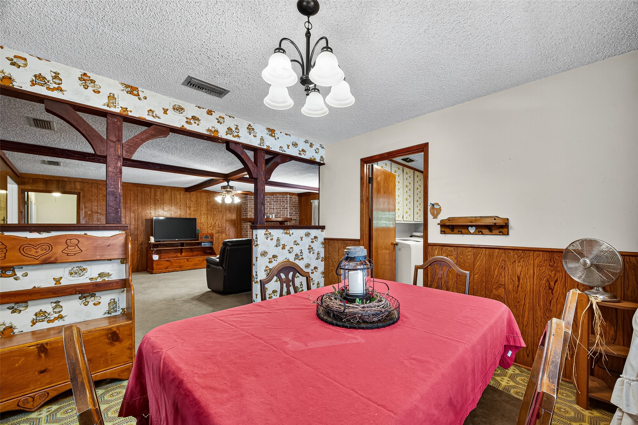 Cozy dining area with vintage charm, featuring wood paneling, patterned wallpaper, and decorative beams. The space is open to a living room with a TV, offering a warm and inviting atmosphere.