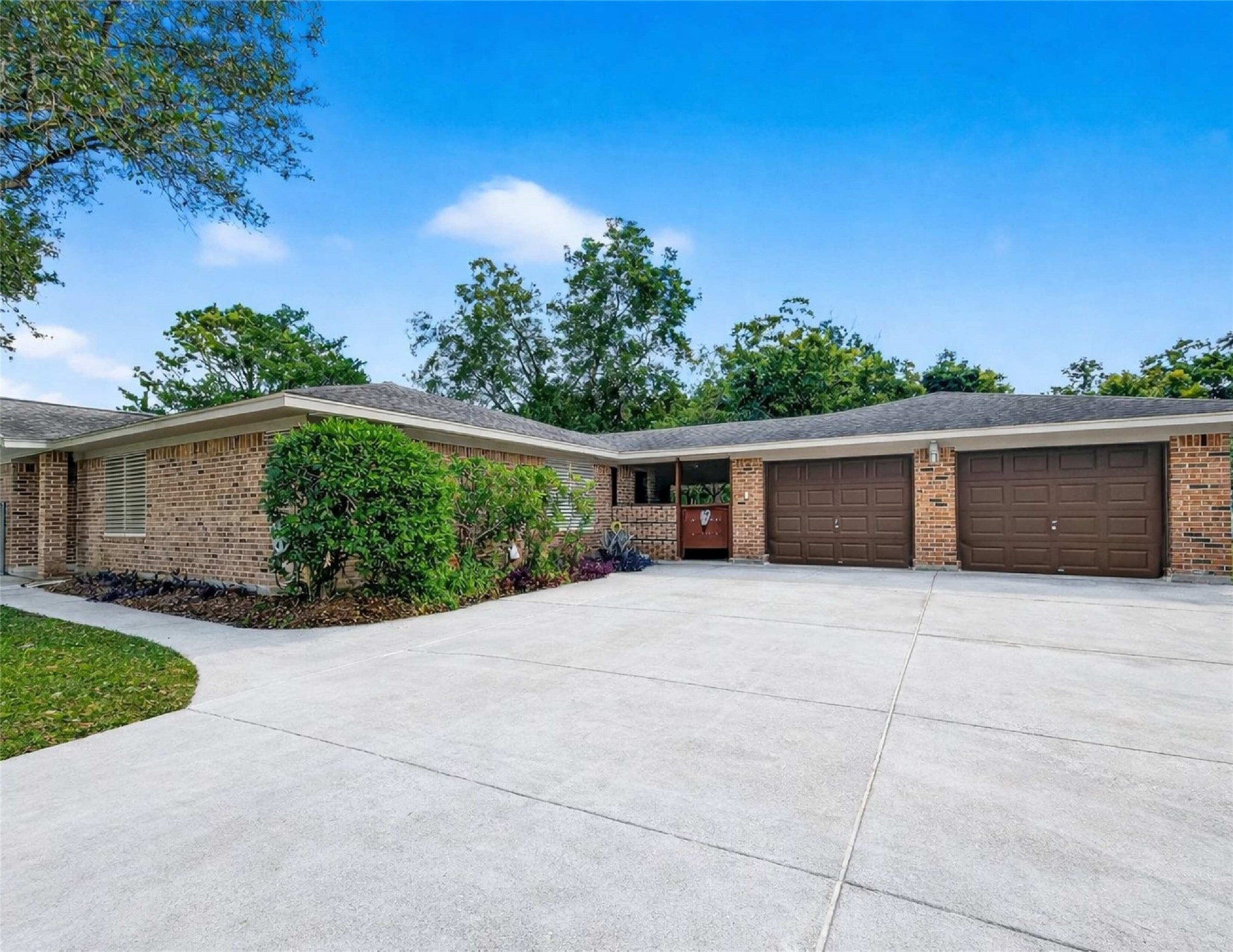 This photo showcases a spacious, single-story brick home with a two-car garage. The property features a large driveway, well-maintained landscaping, and is surrounded by mature trees, providing a serene and inviting atmosphere.