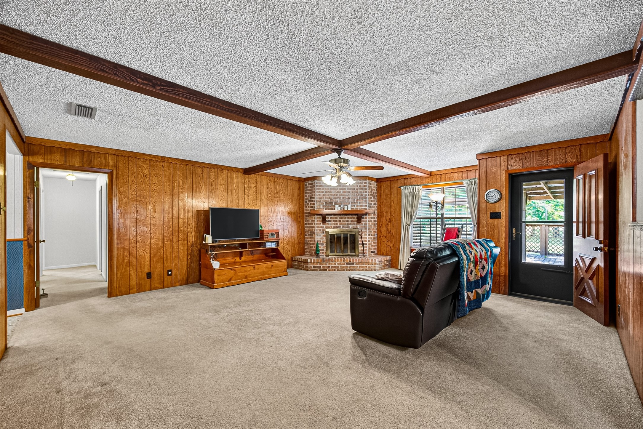 This cozy living room features wood-paneled walls, a brick fireplace, and a ceiling with exposed beams. It has carpeted flooring, a ceiling fan, and large windows allowing natural light. A door leads to an outdoor space.