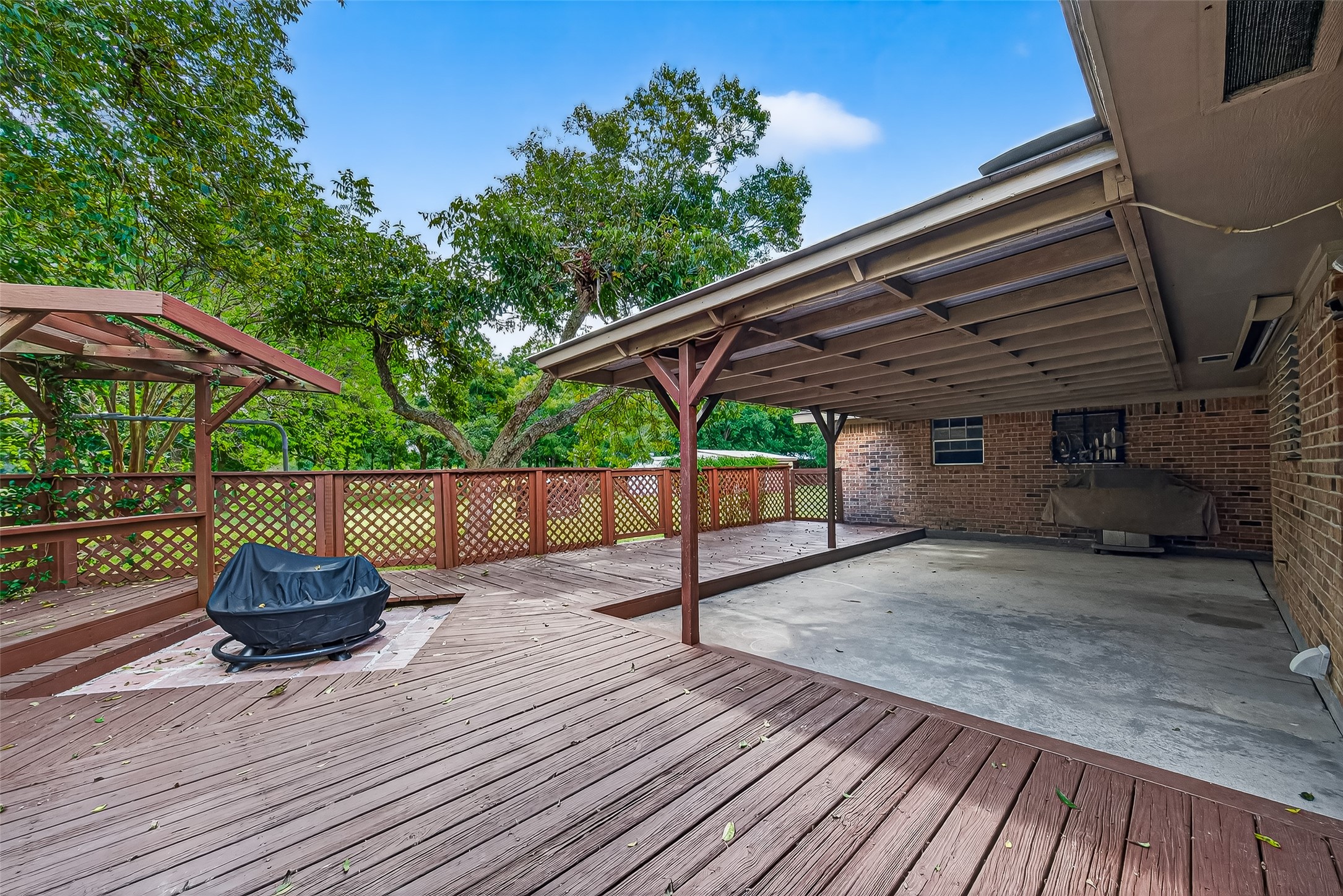 This photo shows a spacious backyard patio featuring a large wooden deck with a fire pit and a covered area for shade and ready for hot tub to be installed. The space is surrounded by lush greenery, providing a private and serene outdoor setting. Perfect for entertaining or relaxing.