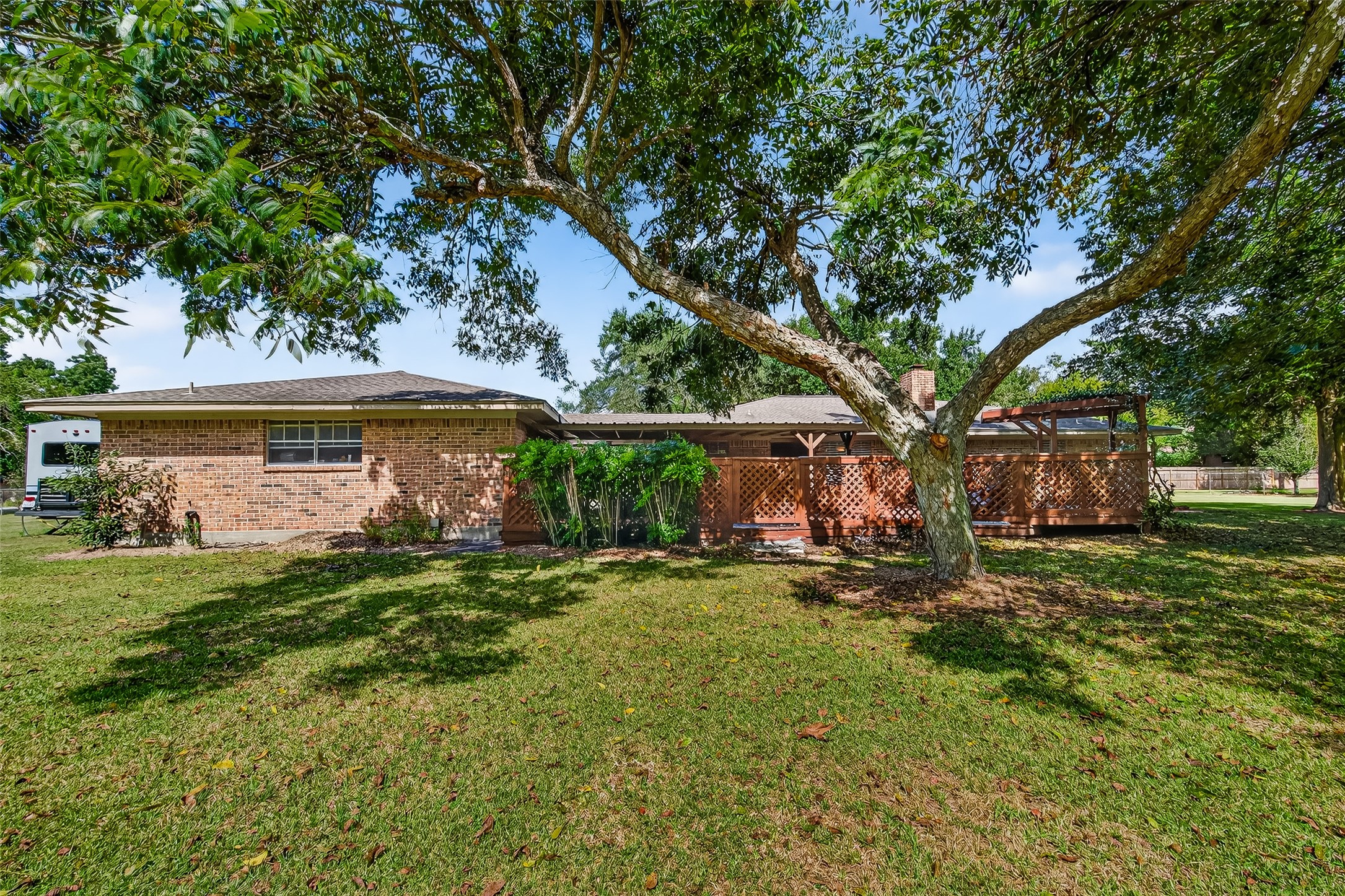Charming brick home with a spacious backyard featuring lush grass and mature trees. A wooden patio area provides shade and outdoor living space, perfect for relaxation and entertaining.