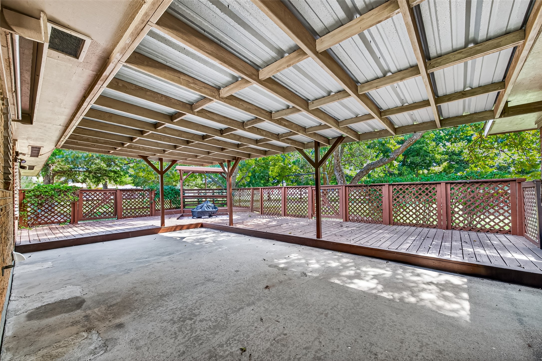 This photo showcases a spacious covered patio with a metal roof and wooden beams. It features a raised wooden deck with lattice railing, offering a private and shaded outdoor area surrounded by greenery, perfect for relaxation or entertaining.