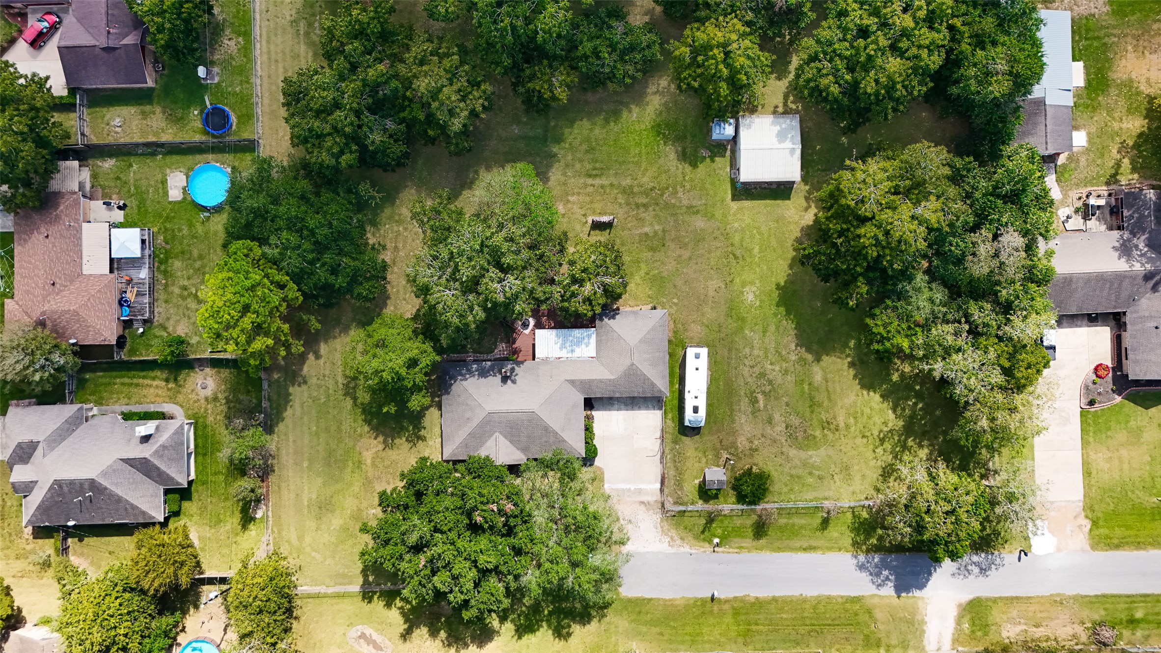 This aerial photo shows a spacious property with a large, well-maintained yard surrounded by mature trees. The house has a sizable driveway and is situated in a quiet, residential neighborhood. There are neighboring homes with pools visible, indicating a family-friendly area.