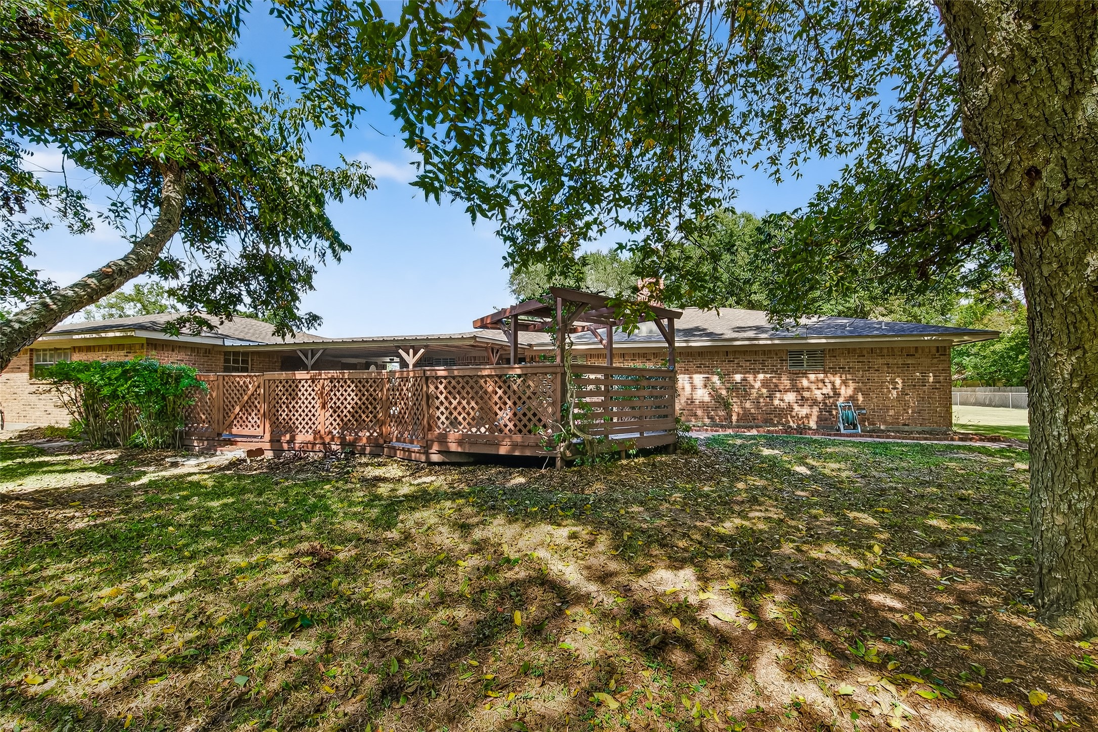 This photo showcases a backyard with a spacious wooden deck and lattice fencing, surrounded by mature trees providing shade. The brick exterior of the house complements the natural setting, creating a cozy and inviting outdoor space.