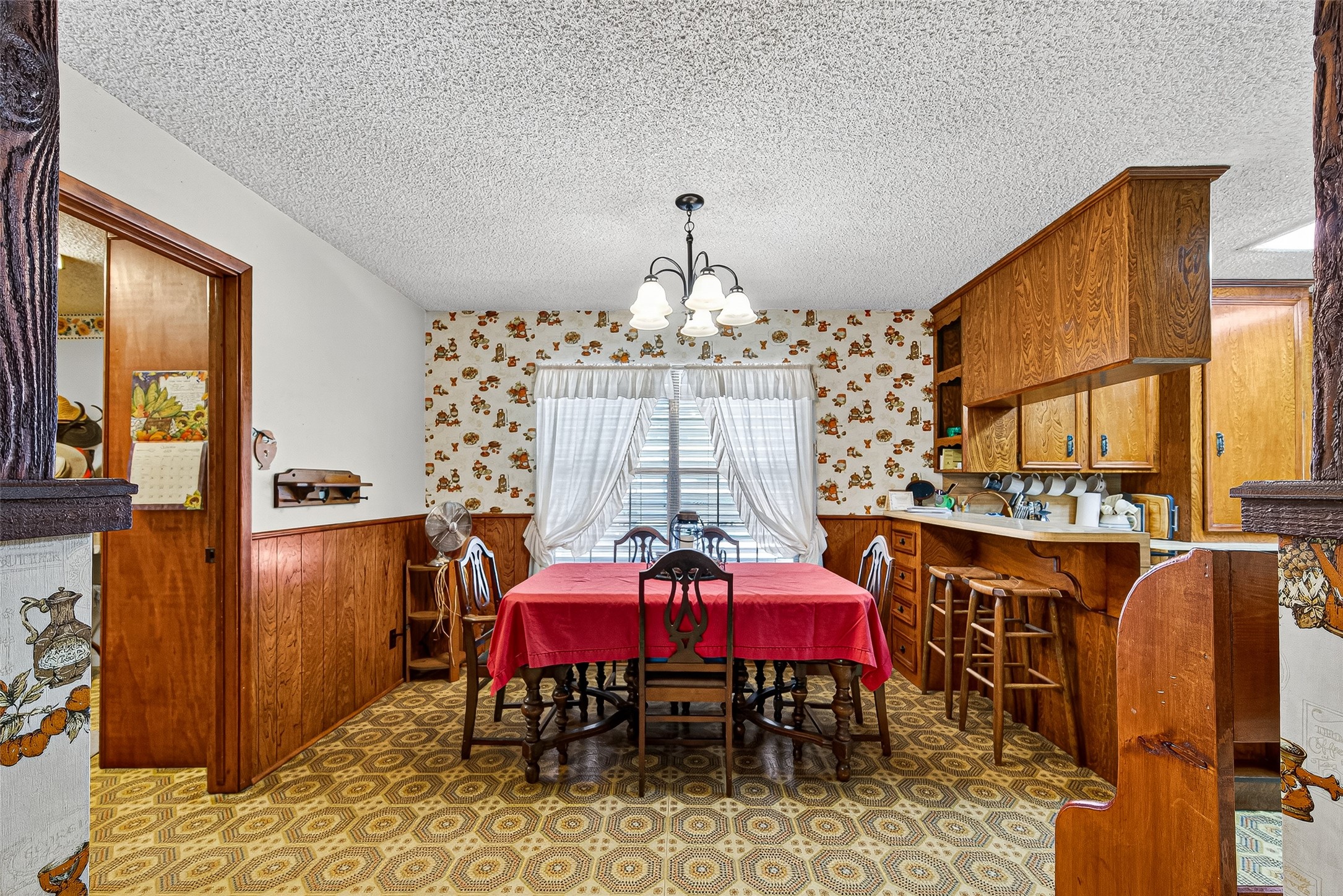 This cozy dining area features vintage charm with floral wallpaper and wood paneling A built-in kitchen nook with ample storage and counter space. A window with drapes allows natural light to brighten the space.