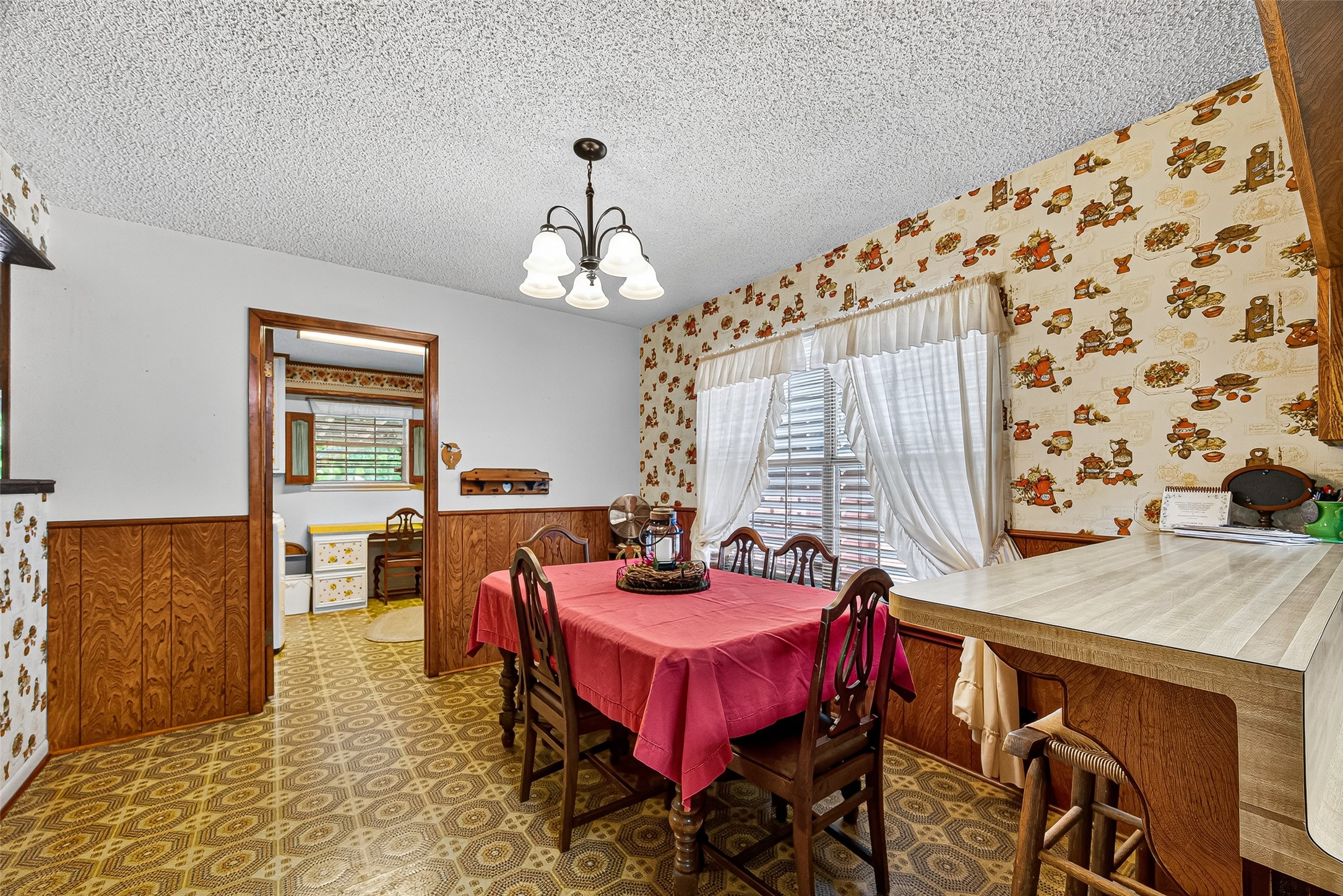 This cozy dining area features vintage wallpaper and wood paneling, with a bright window and overhead lighting. The space connects to a kitchen through a doorway, and includes a practical breakfast bar for additional seating.