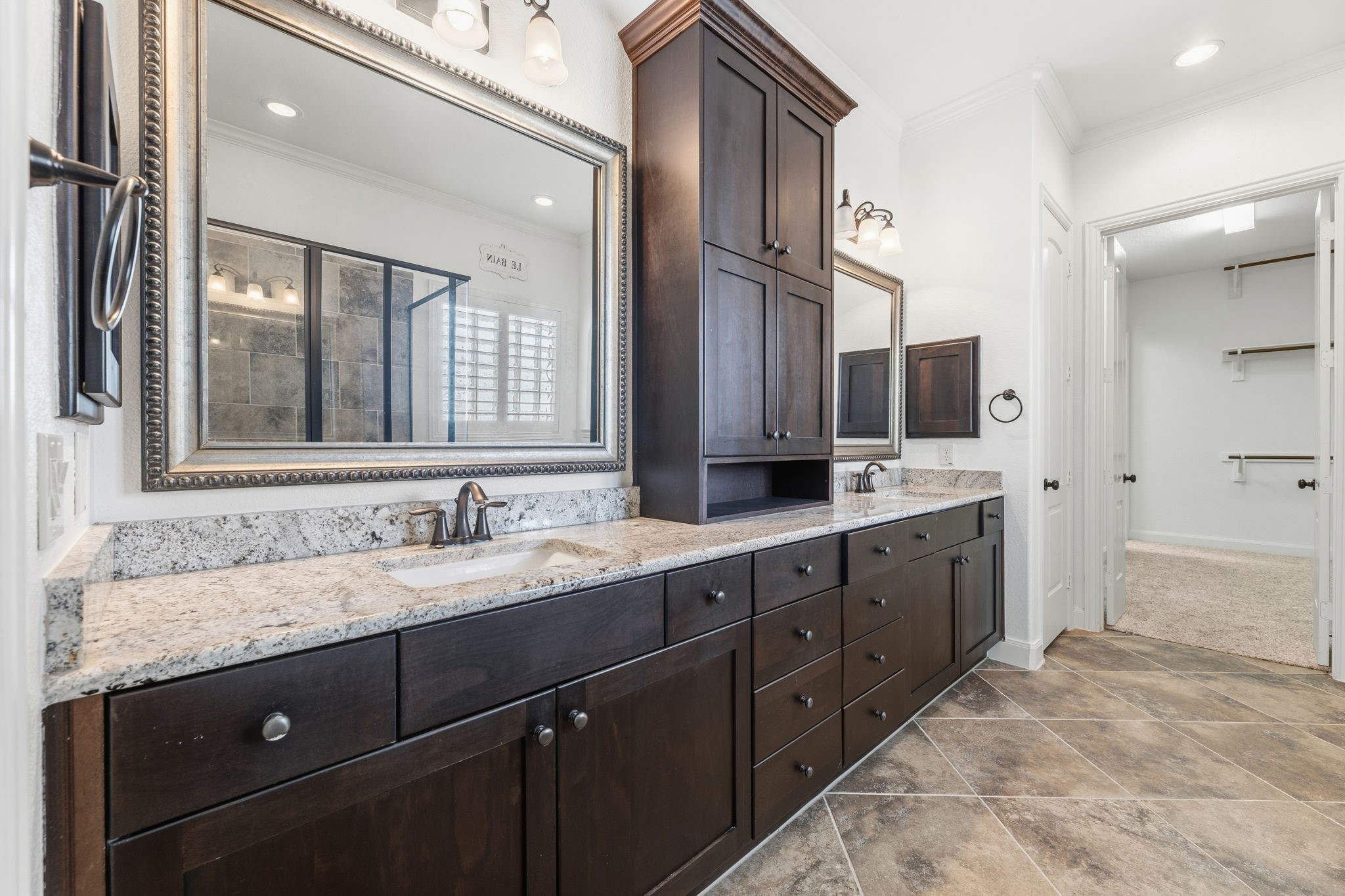 Primary bath featuring granite countertops, dark wood cabinetry, dual sinks, and a spacious layout that connects to a walk-in closet.