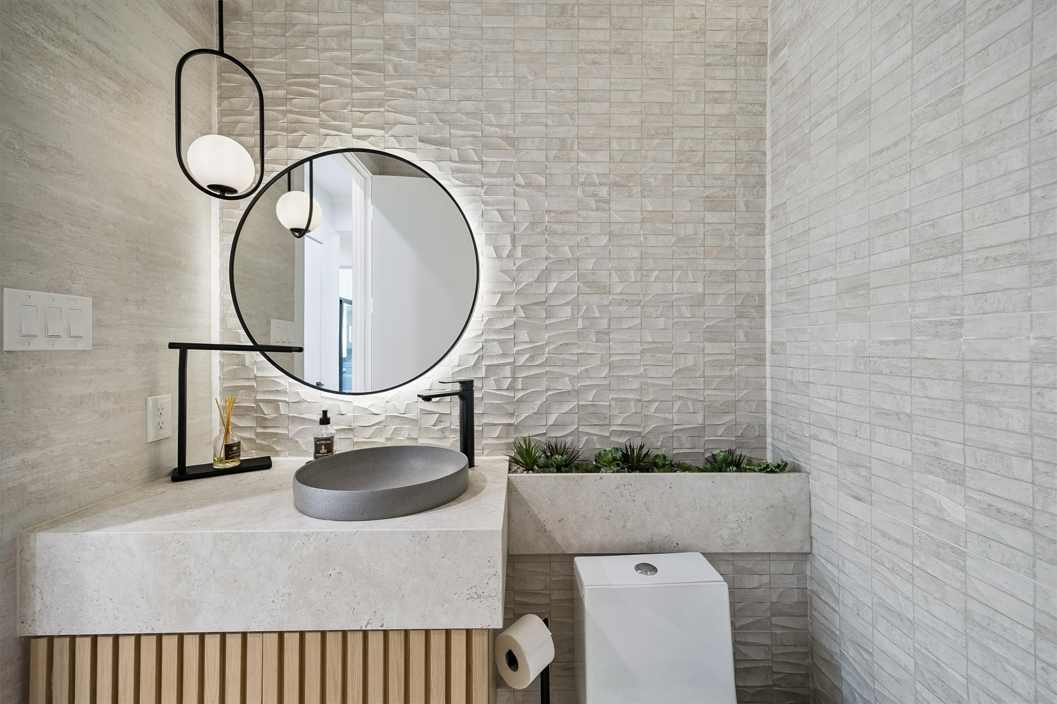 A beautifully curated powder room makes a memorable statement with textured finishes, a sculptural vessel sink, contemporary fixtures, and a distinctive illuminated mirror.