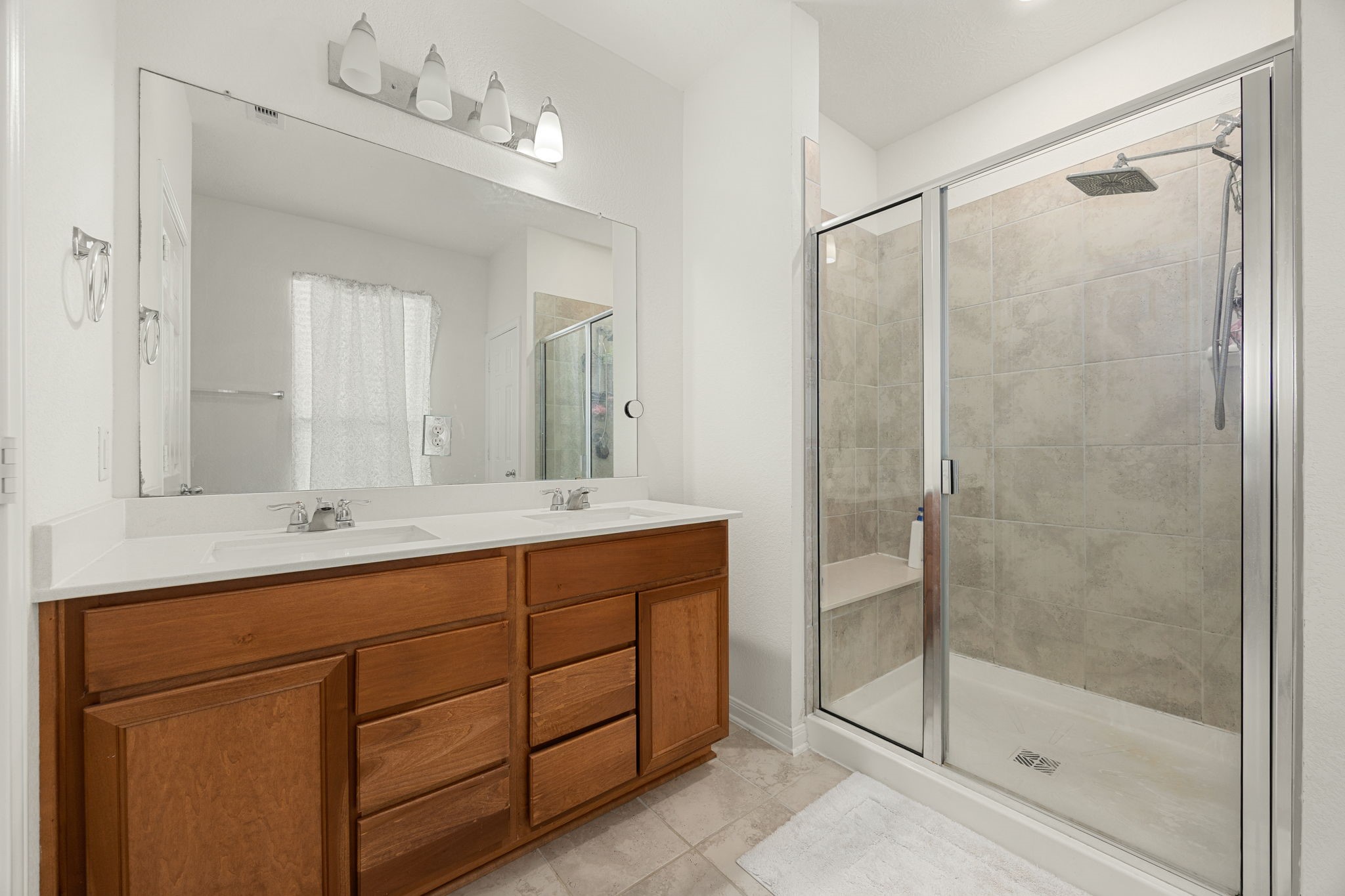 Pamper yourself in the well-appointed primary bathroom, highlighting excellent storage capacity and clean finishes. The oversized step-in shower boasts a premium rain-style showerhead and dedicated seating, beautifully complementing the long wood-toned vanity with two sinks, durable tile flooring, and bright, high-ceiling layout.