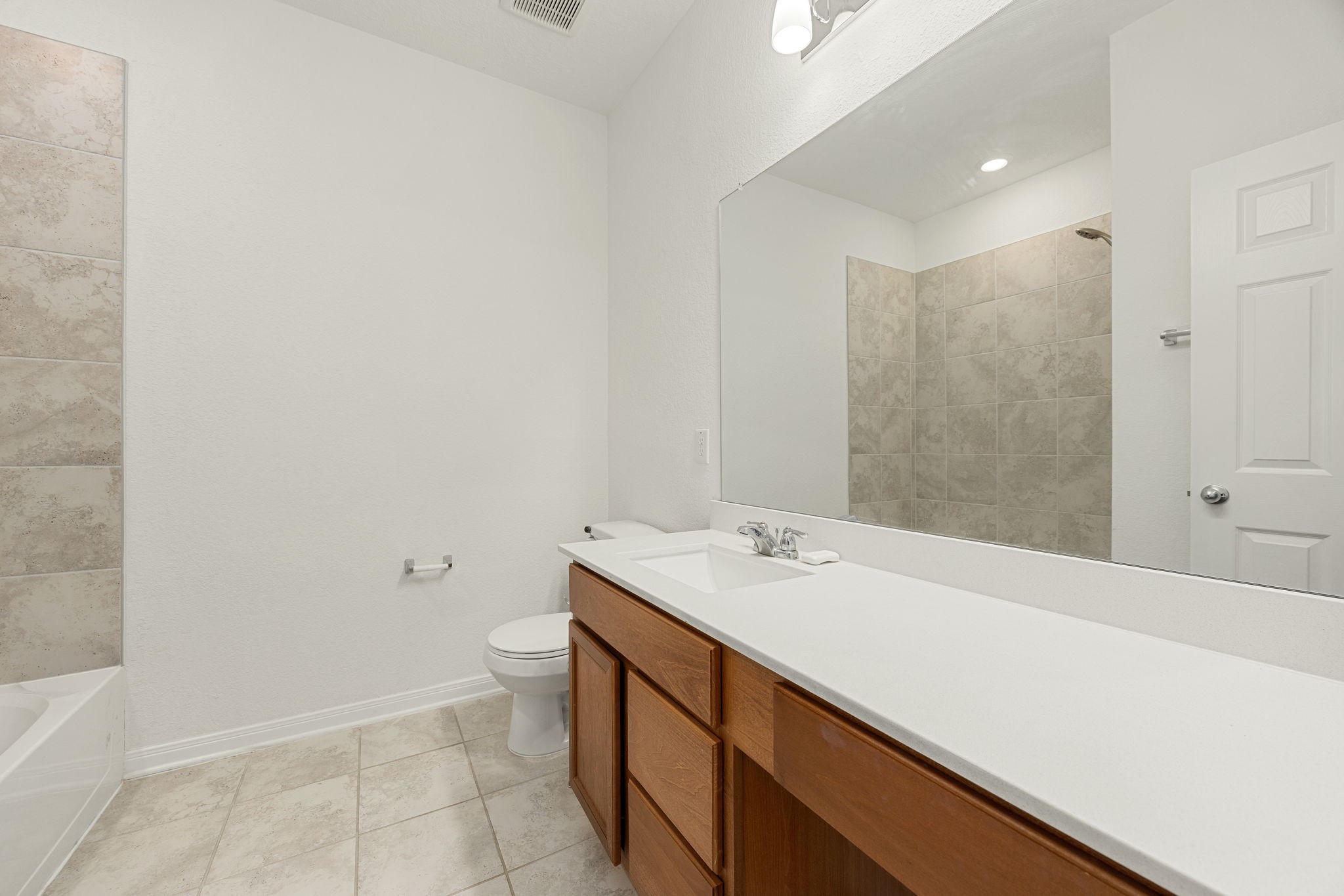 The spacious secondary bathroom features an expansive vanity with an integrated sink, an extra-wide mirror, and a convenient sit-down vanity knee space. The space is complete with neutral tile flooring and a clean shower/tub combo wrapped in classic oversized tiling.