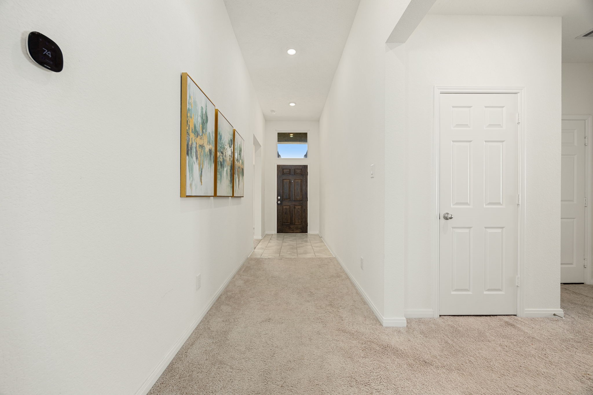 A view looking back toward the front entry highlights the home's impressive high ceilings and spacious layout. This long, architectural hallway features neutral plush carpeting, modern recessed lighting, and crisp white walls that provide a bright and open atmosphere from the moment you step inside.