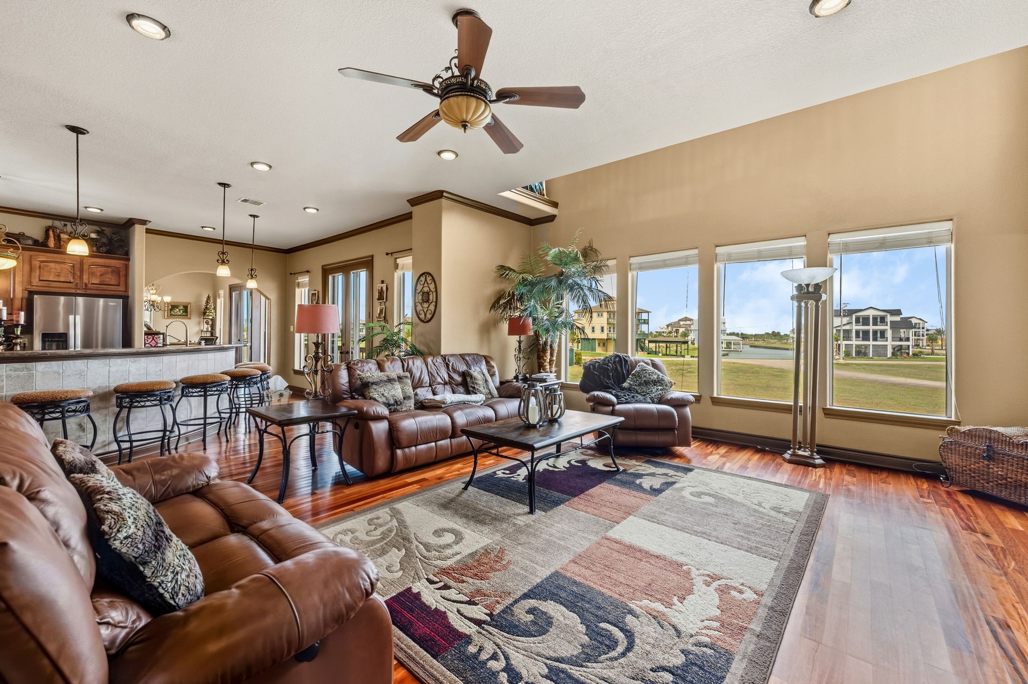 Spacious living area with rich wood flooring and large windows offering scenic views. The open layout seamlessly connects to a modern kitchen with bar seating, creating an inviting space for relaxation and entertaining.
