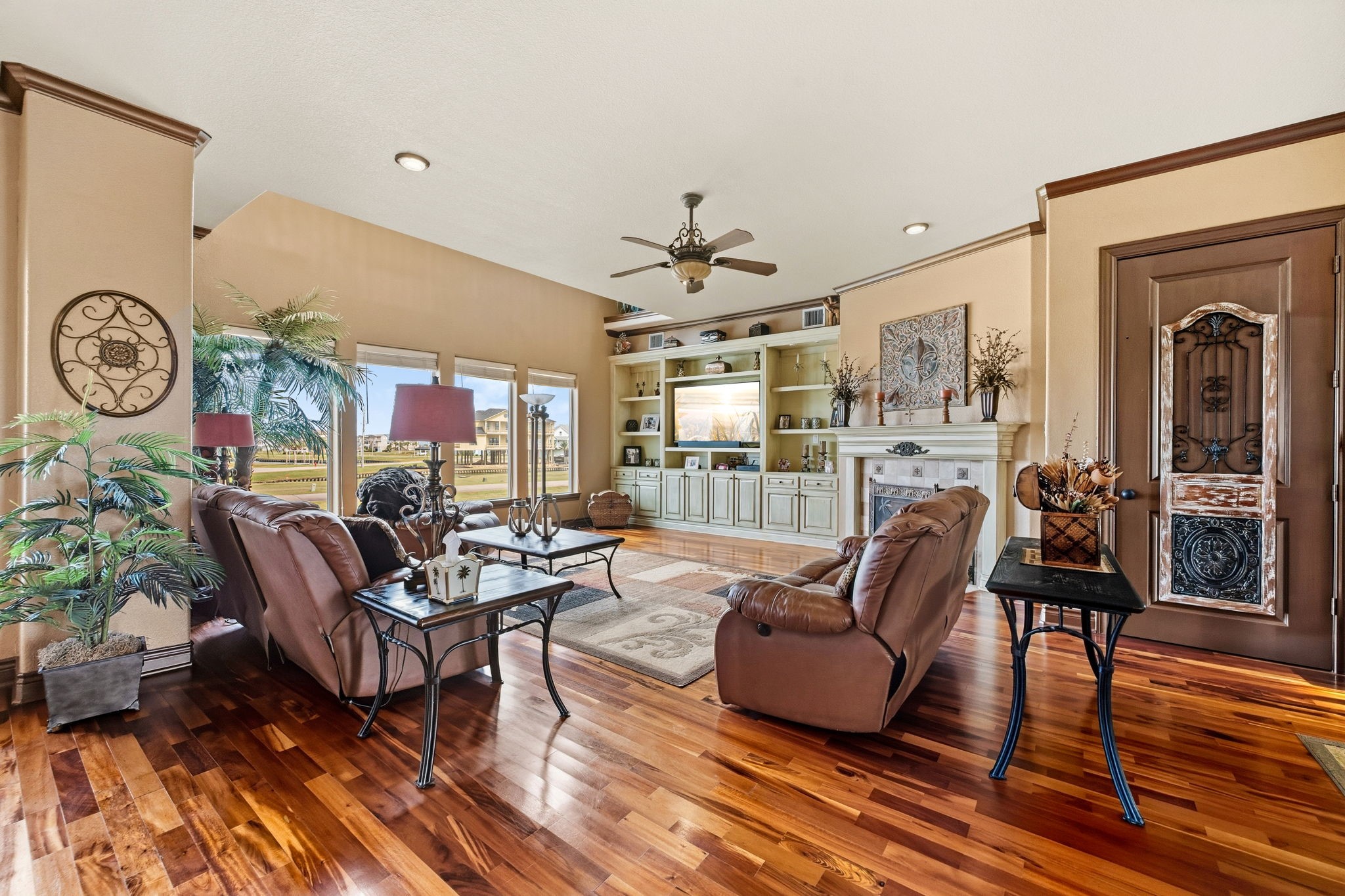 Spacious living room with elegant hardwood floors, cozy leather seating, and a built-in entertainment center. Large windows offer abundant natural light and picturesque views, complemented by tasteful decor and a stylish ceiling fan.