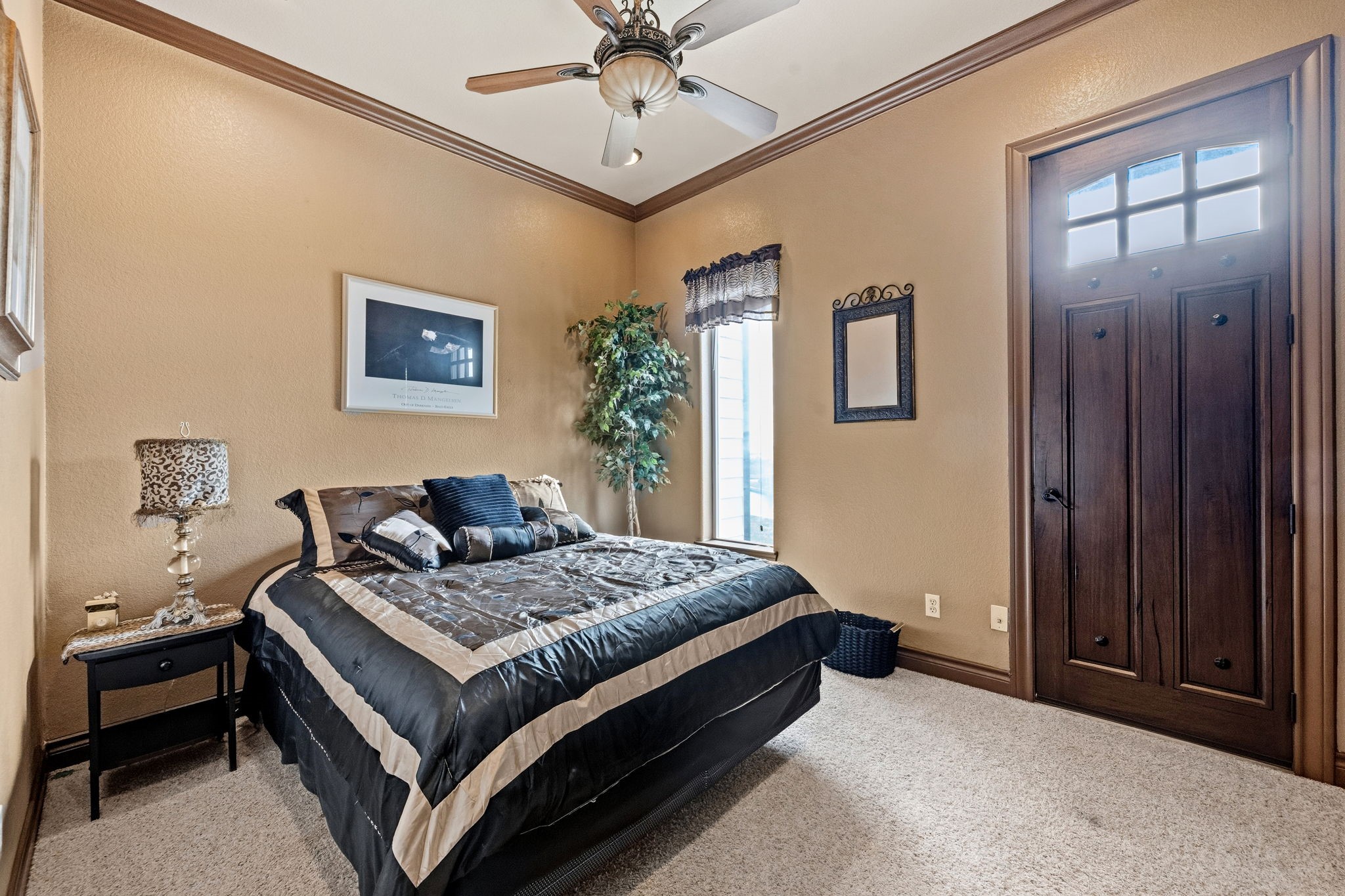 2nd bedroom with warm tones, featuring a ceiling fan, plush carpeting, and a window for natural light. A rich wooden door adds a touch of elegance.