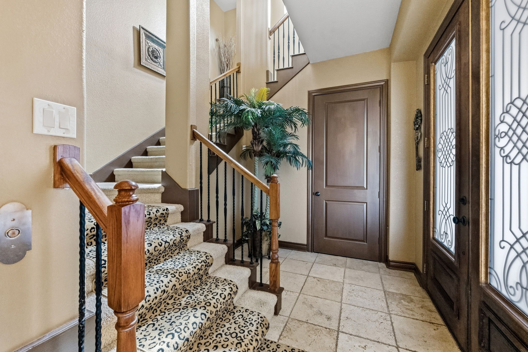 Elegant entryway featuring a carpeted staircase with a bold pattern, intricate ironwork on the doors, and warm wood accents complemented by tile flooring.