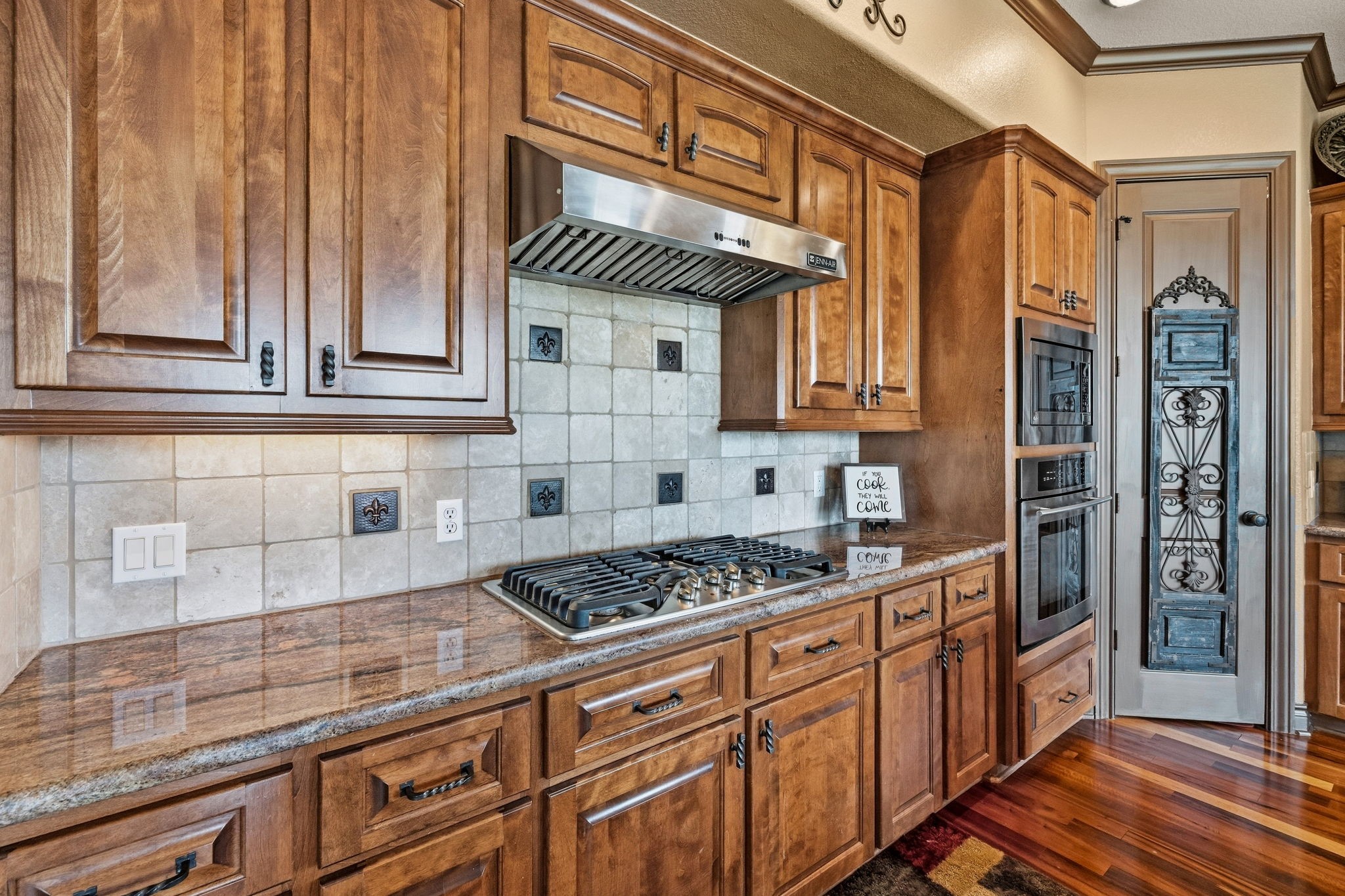 Elegant kitchen featuring rich wood cabinetry, a sleek stainless steel range hood, and granite countertops. The tile backsplash and double ovens add a touch of sophistication, while a decorative pantry door completes the space.