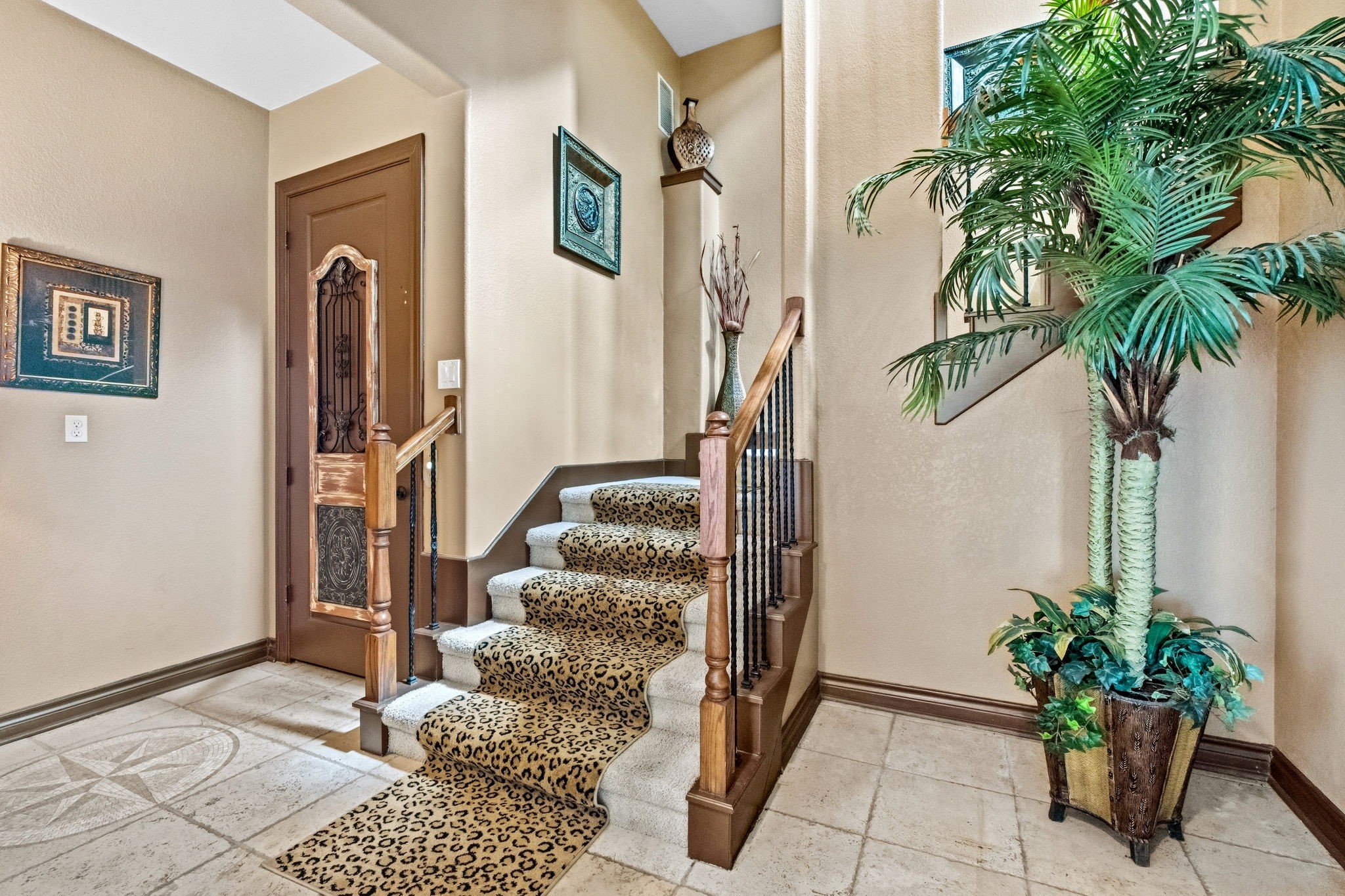 Elegant staircase with ornate wooden railing and leopard-print carpet, complemented by decorative wall art and a lush potted plant, creating a welcoming entrance.