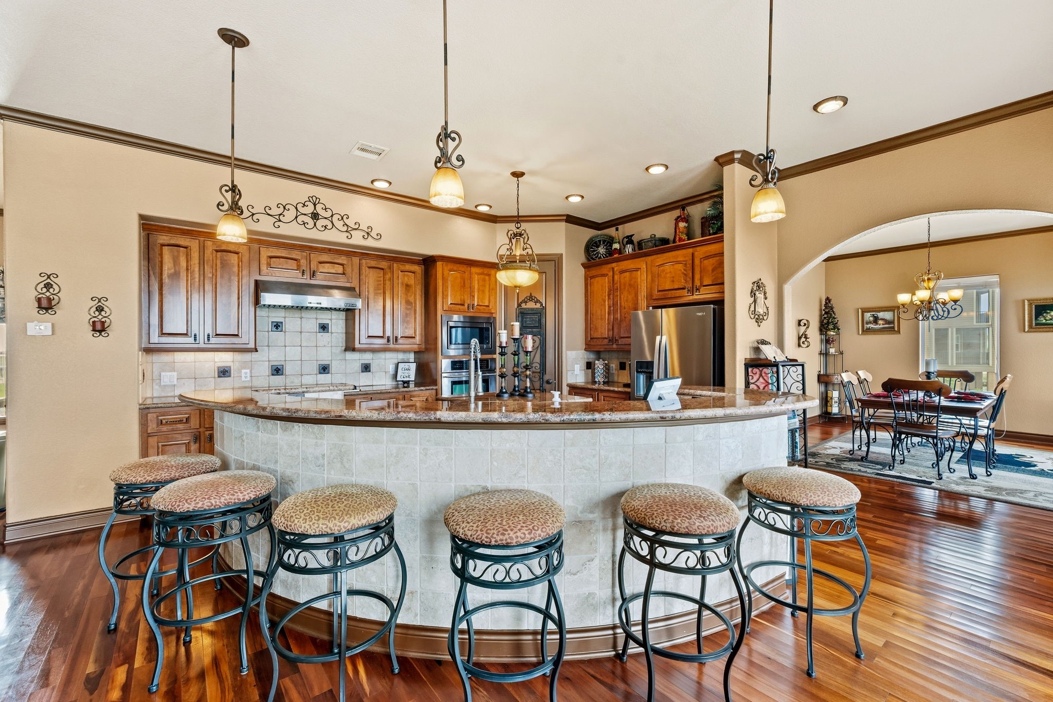 Spacious kitchen with rich wood cabinetry, a large curved island with seating, and elegant pendant lighting, seamlessly connecting to a cozy dining area.