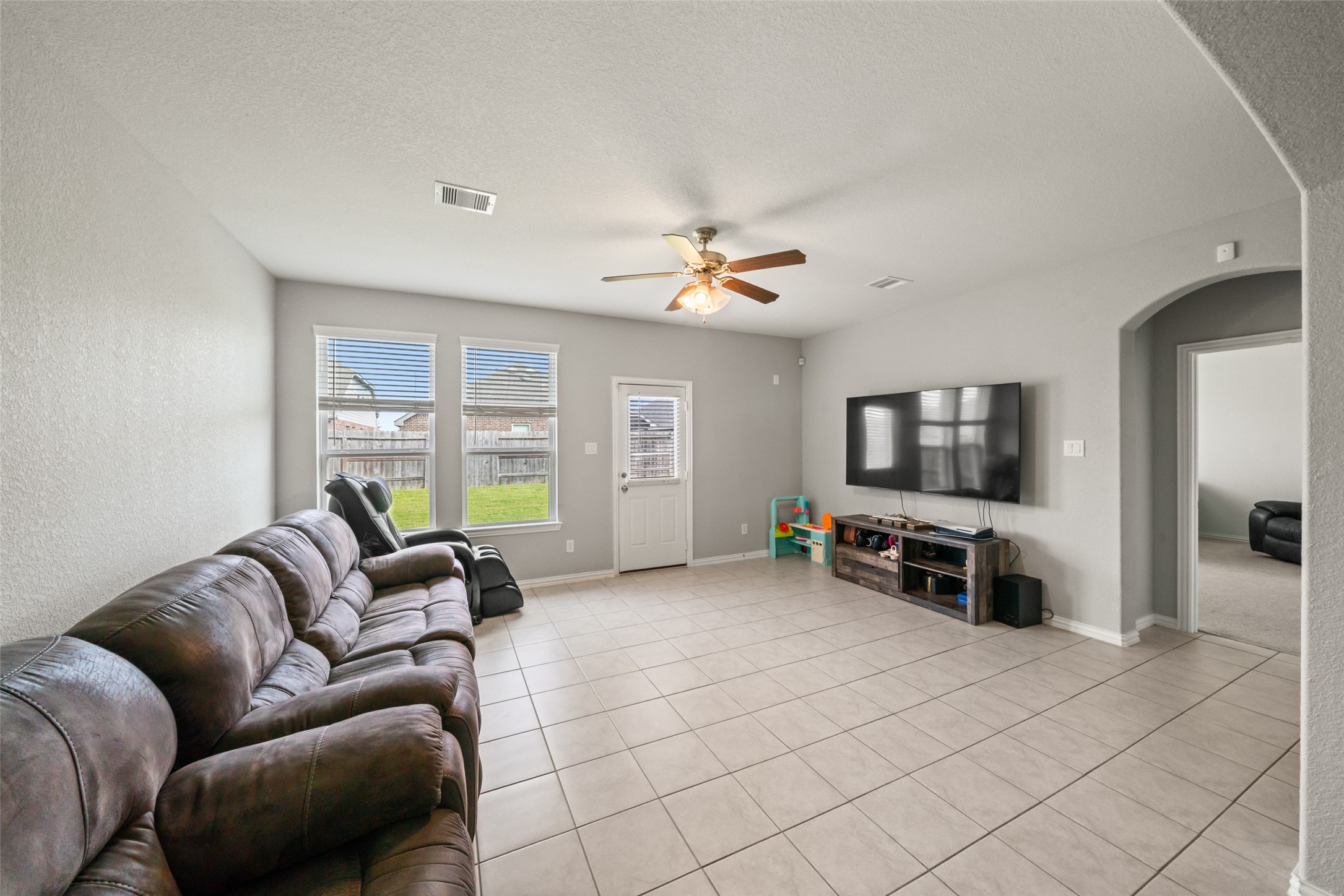 Spacious living room with a tiled floor, featuring a cozy reclining sofa and large windows offering plenty of natural light.