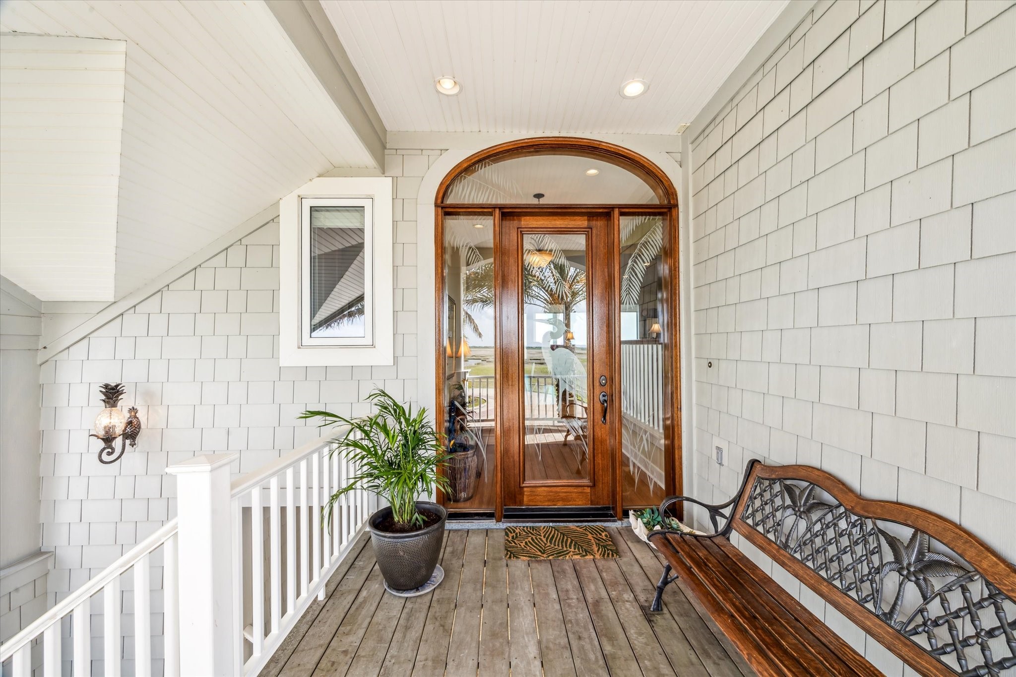 Welcome guests in style with this inviting front entry, featuring rich wood-framed etched glass door beneath an elegant arched transom. The covered porch is accented with low-maintenance Hardie shingle siding, warm-toned IPE decking, and coastal-inspired details including a decorative wall sconce and classic white railing, making this space the perfect transition into the relaxed yet refined interior of the home