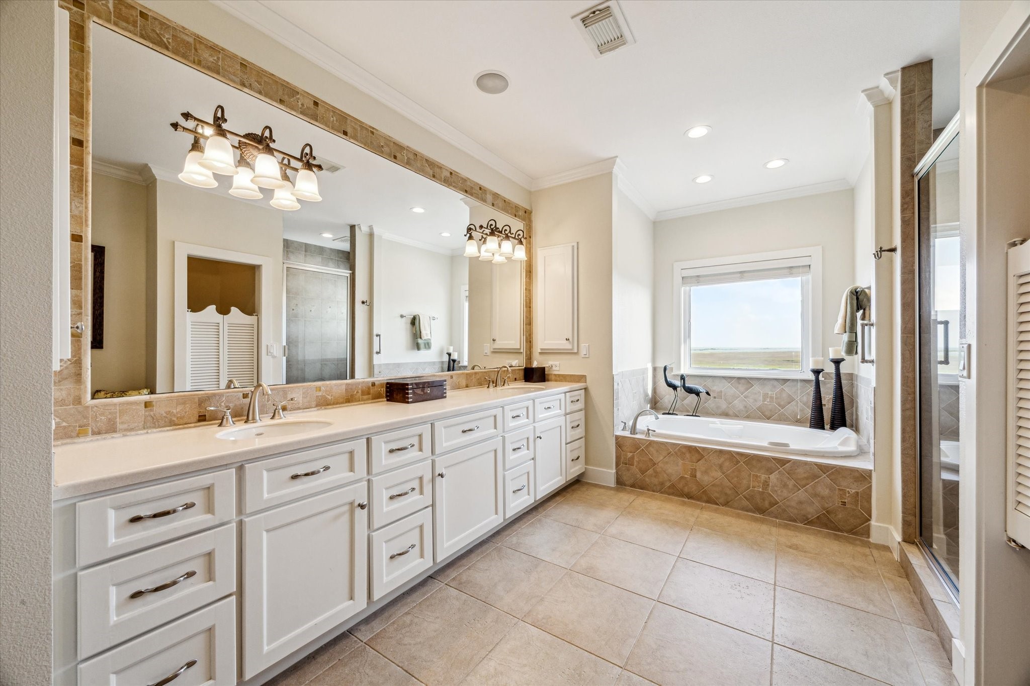 This elegant bathroom features a spacious, spa-like design with a large dual vanity topped with sleek countertops and framed by a full-width mirror with decorative lighting. The custom cabinetry offers abundant storage with a clean, white finish that complements the neutral tile flooring.