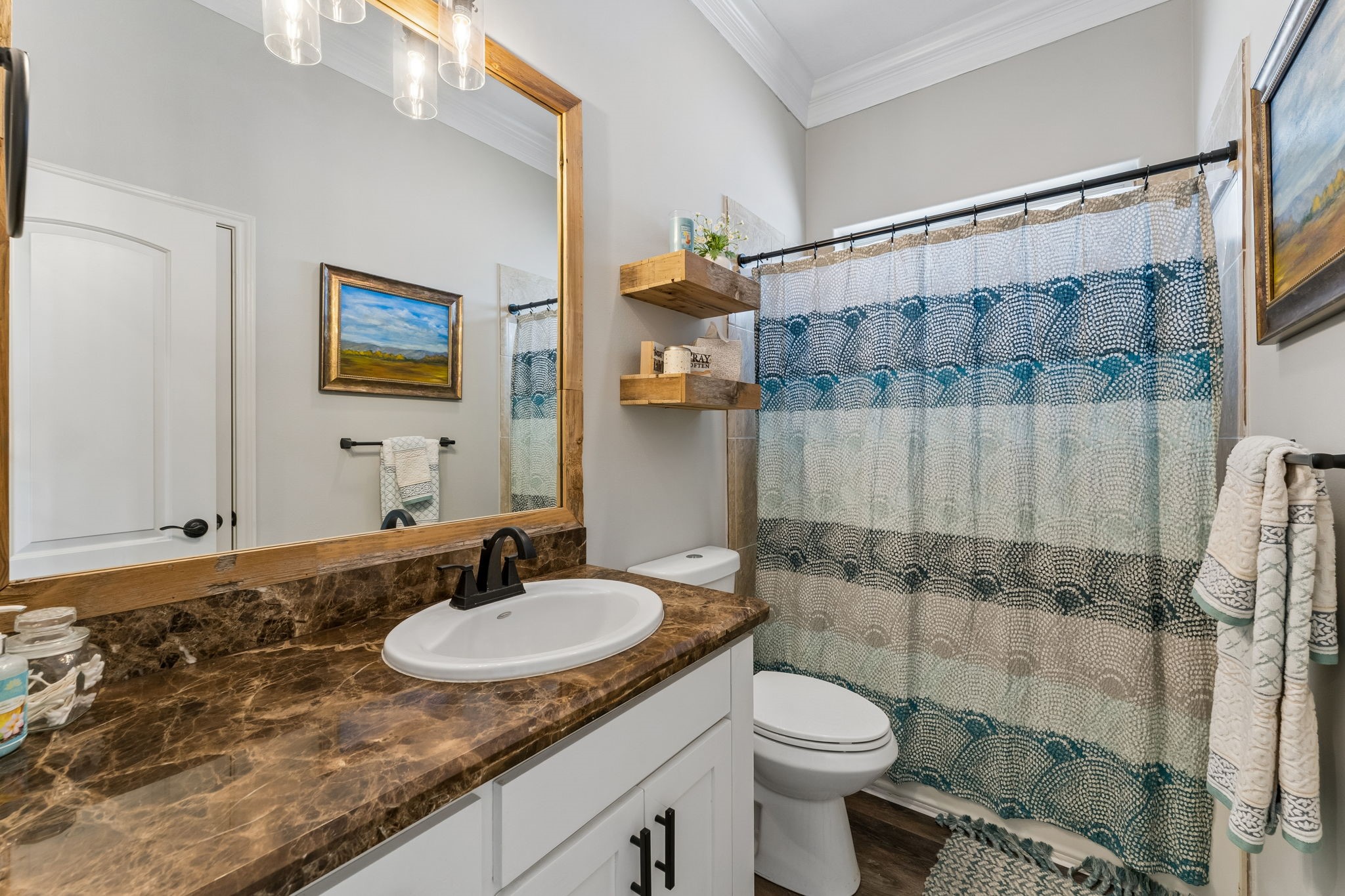 Full bath features a spacious vanity with rich stone countertops and a large framed mirror. Floating wood shelves add warmth and convenient storage.