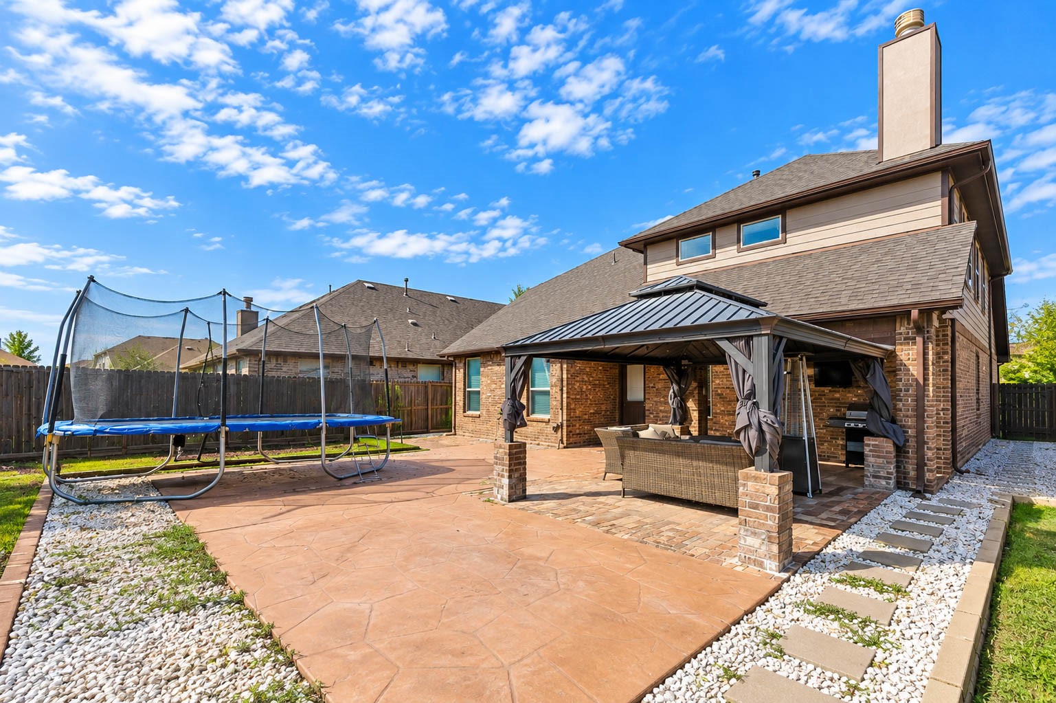 A view of the back of the house shows a two-story brick elevation with a chimney, a spacious stamped concrete patio and a covered gazebo with comfortable seating. The yard has room for kids to play and adults to gather. A wood fence provides privacy. Large windows on the back of the house bring daylight inside and give you a view of the yard from the living areas. It makes the whole backyard feel like an extension of the home.