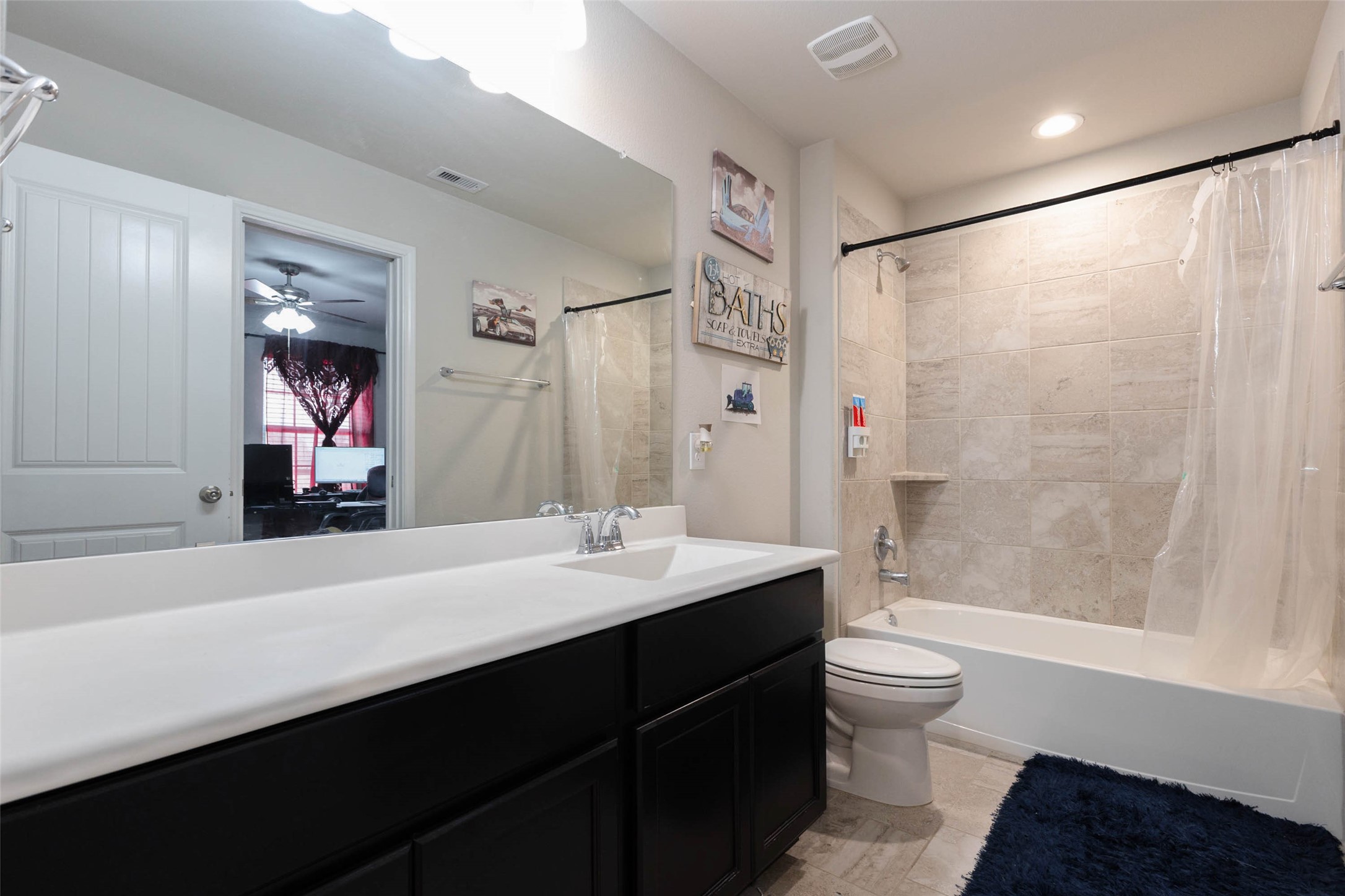A full bathroom with a long vanity, a mirrored wall and a tub and shower combination with a tile surround and curtain. The vanity has dark cabinetry with a white countertop, which gives you plenty of storage and counter space. A door on the left opens to a bedroom. This bath serves the secondary bedrooms well and keeps morning routines on schedule for a busy household. It is a sensible and comfortable space for family and guests.