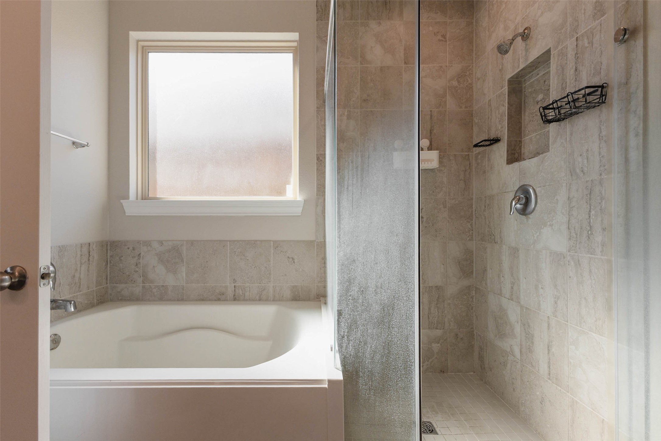 A soaking tub sits beside a frosted window and a glass-enclosed shower with a tiled surround and a built-in niche. Warm, natural-toned tile covers the shower walls and the tub deck, which gives the room a spa-like quality. The separate tub and shower let you choose between a long soak and a quick rinse. It is a peaceful, private space at the end of the day.