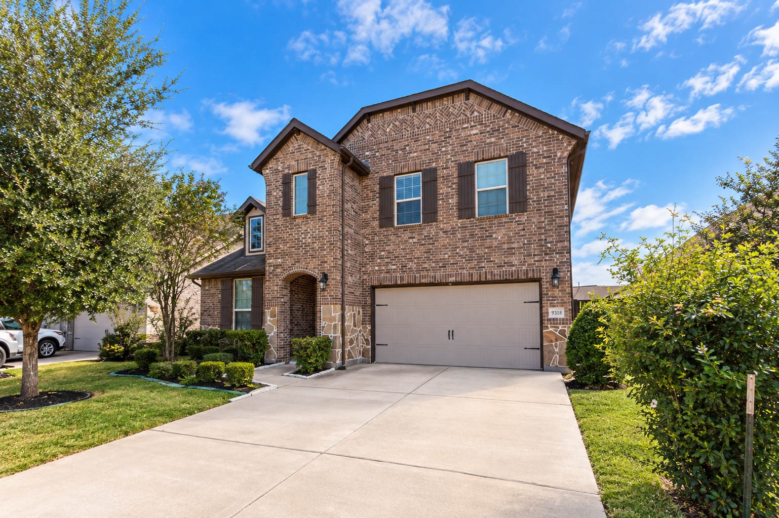 Another angle of the front shows the full driveway, the two-story brick exterior and the way the roofline steps back above the garage. Shrubs along the right side and a mature tree on the left frame the home on both sides. The lot has room for cars and guests to park without crowding the street. Curb appeal like this sets the tone for what you will find inside. It is a welcoming first look at the home from every direction.