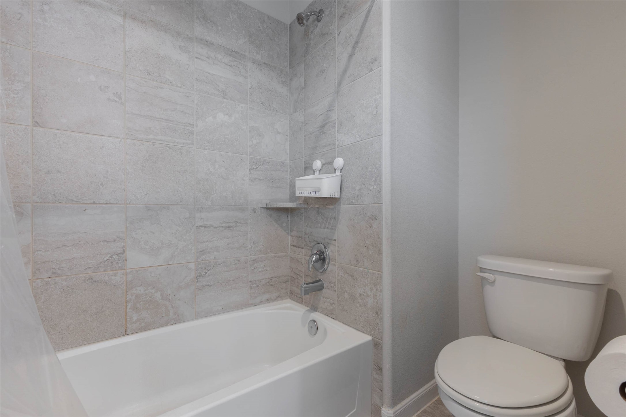 A tub and shower combination with a tiled surround, a soap shelf and a toilet beside it. Large gray tiles run floor to ceiling around the tub, which makes cleaning easy and gives the room a modern feel. A window lets in light, and the bath is conveniently located near the bedrooms. This is a practical bathroom for kids, guests or anyone who prefers a classic bath and shower setup. Easy to keep clean and a comfortable fit for the whole family.