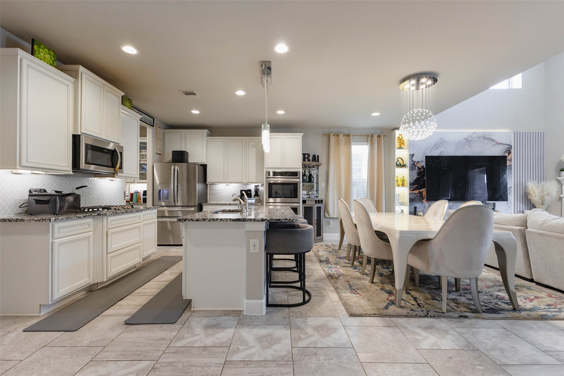From the end of the kitchen the sight line stretches across the island to the dining table and the family room with its large TV wall. Stainless steel appliances, a stainless refrigerator and a double-width run of cabinets give the cook everything within reach. Tall windows on the far side let in daylight and a crystal chandelier hangs over the dining area. The open plan keeps conversation flowing from one space to the next.