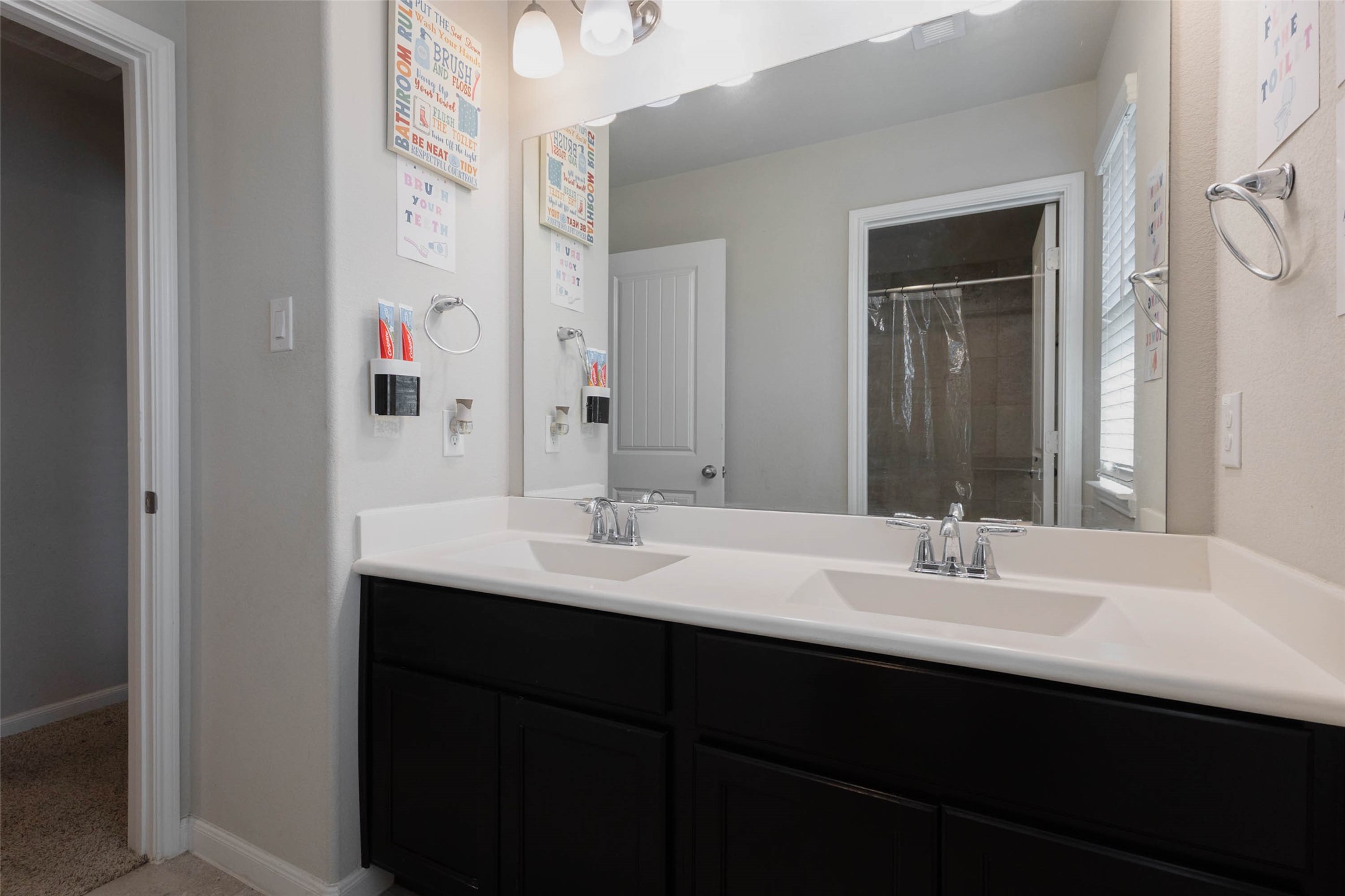 A bathroom with a long dual-sink vanity, dark cabinetry and a white countertop, plus a large mirror with a lighted fixture above. A shower enclosure sits behind the mirror and a doorway opens to the hall. Two sinks help busy mornings run smoothly, and the dark cabinets provide plenty of storage underneath. The contrast between dark and light finishes gives the space a modern look. Everything is within easy reach in this well-planned space.