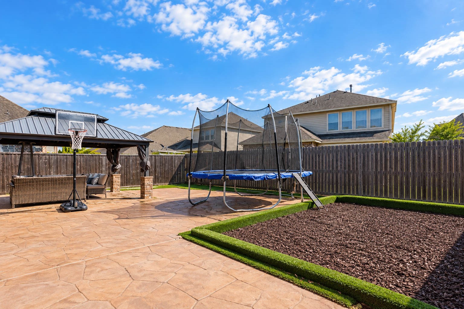 Looking across the backyard toward the gazebo, you see the stamped concrete patio, artificial turf, a bed of fresh mulch edged with clipped hedges and a basketball goal. The wood fence wraps the yard, and the sky gives the scene a bright, open feel. Low-maintenance landscaping means more time to enjoy the space and less time working on it. A great setting for fun and relaxation. Plenty of reasons to spend the weekend outside.