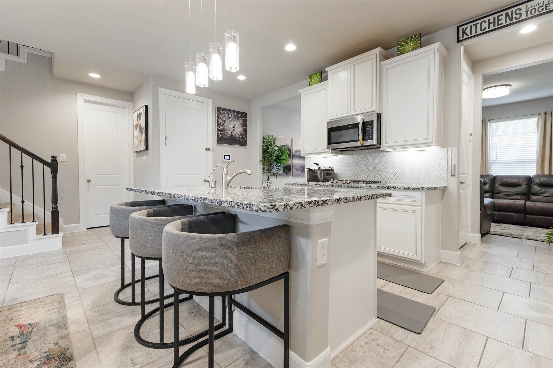 The kitchen island has a granite top, three bar stools and pendant lights hanging above, perfect for casual meals and conversation. White cabinets, a stainless microwave and under-cabinet lighting give the room a bright, clean look. A pantry door and a staircase sit near the kitchen, which keeps storage close and traffic flowing. The seating at the island lets guests visit with the cook without crowding the work zone.