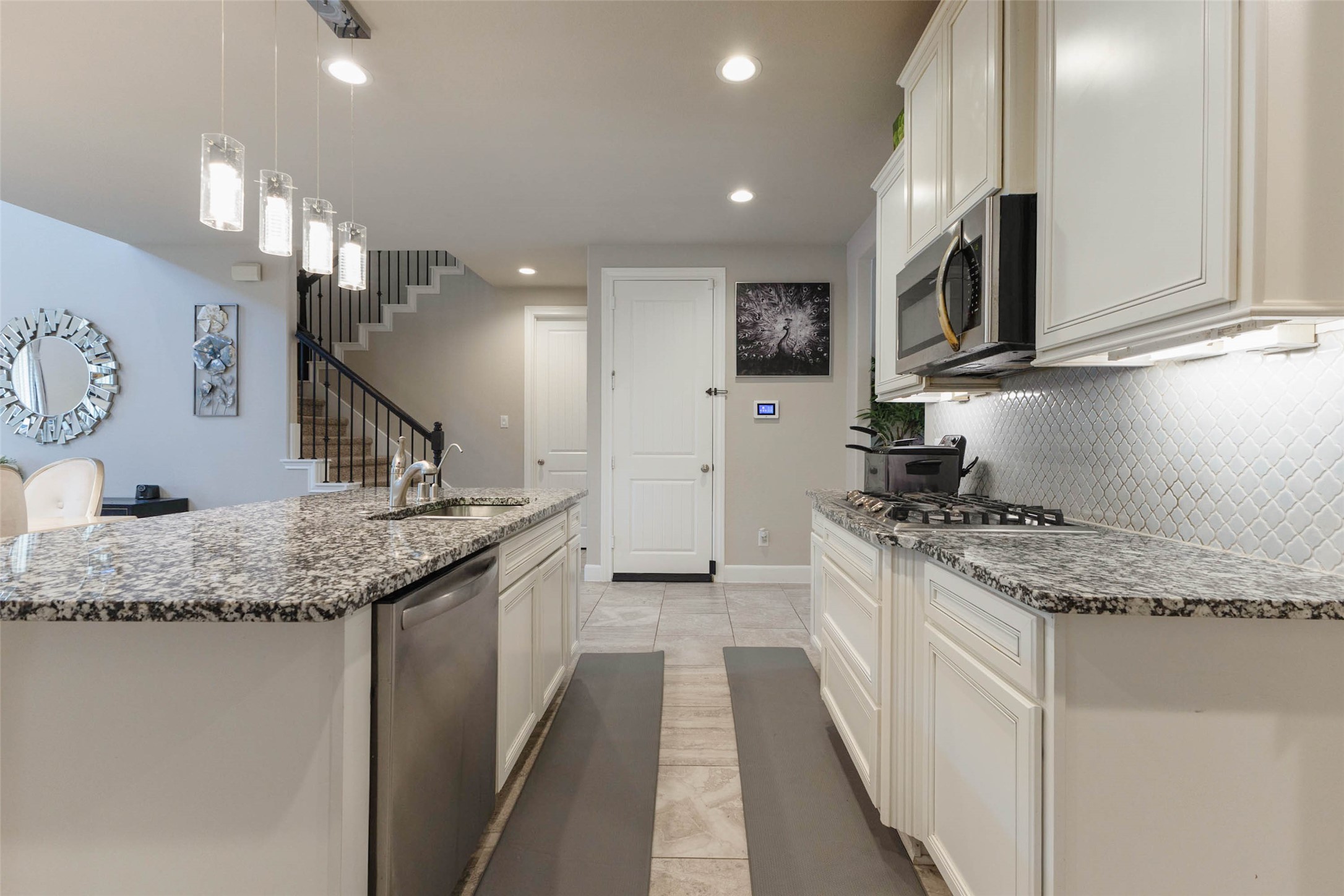 The kitchen includes a cooktop with a decorative tile backsplash, a stainless dishwasher beside the sink and granite counters running the full length. Pendant lights above the island, recessed lighting overhead and white cabinets reaching the ceiling make the room feel bright. A staircase and a closet door appear in the background, a handy spot for storage. Everything in this kitchen is built to handle daily use and weekend hosting.