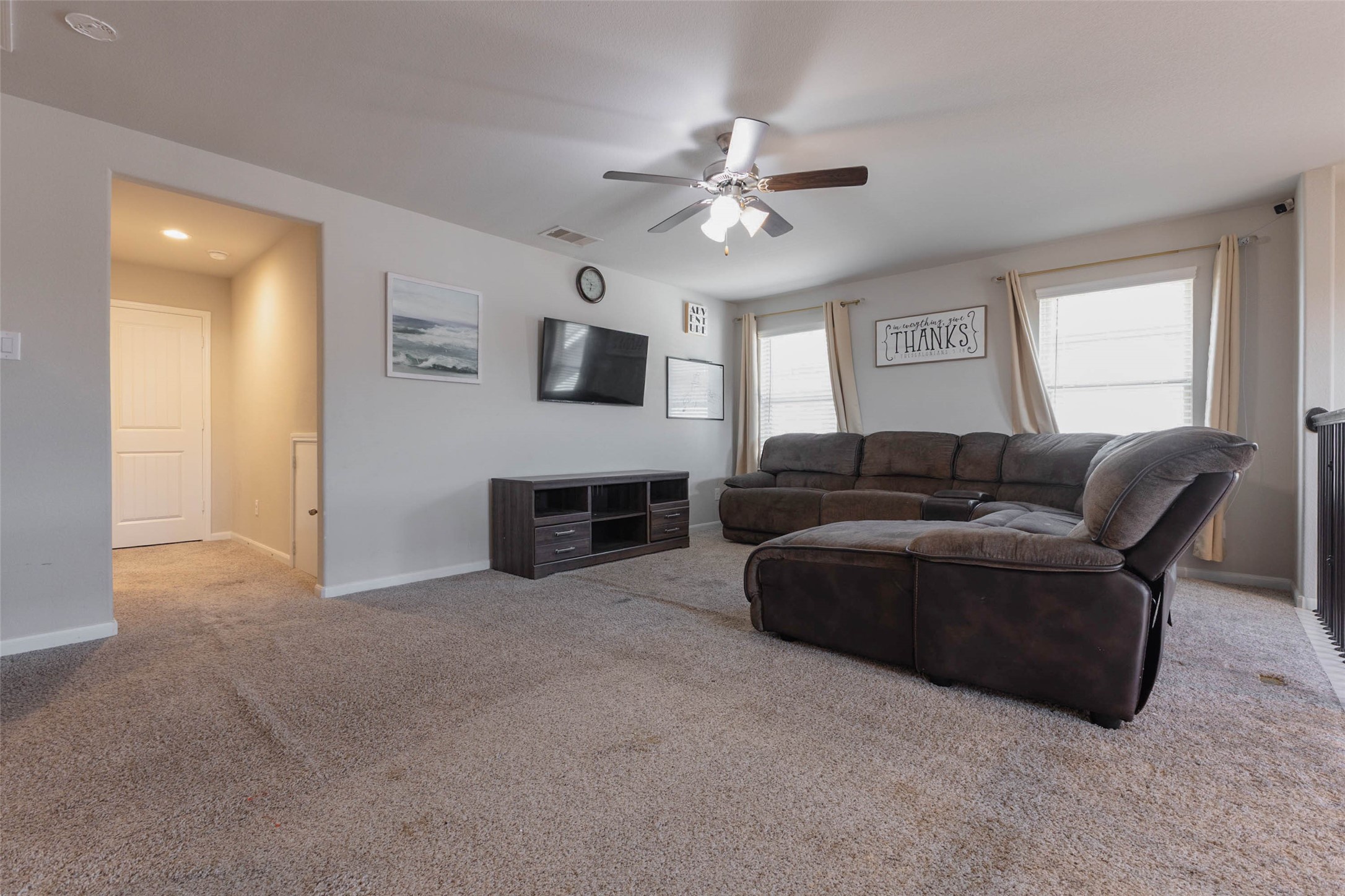 Another angle of the upstairs game room, showing a TV wall, a deep sectional and a doorway leading to the hall and bedrooms. The room is bright, spacious and flexible, with enough floor space for game night, a play area or a workout zone. A ceiling fan and large windows keep the room comfortable. With the railing on one side, the room feels open and connected to the main living space below.