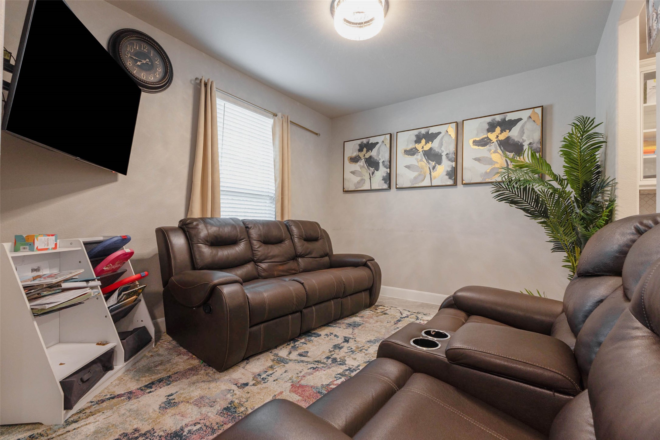 A second living space with a wall-mounted TV, a leather sofa and a pair of recliners, set in a room with soft neutral walls, a ceiling light and a large window. The room is flexible enough to serve as a media room, a den or a play space. Built-in shelving on the right edge adds storage for books and games. It gives the family a place to relax away from the main living area. Comfortable for movie nights and quiet afternoons alike.