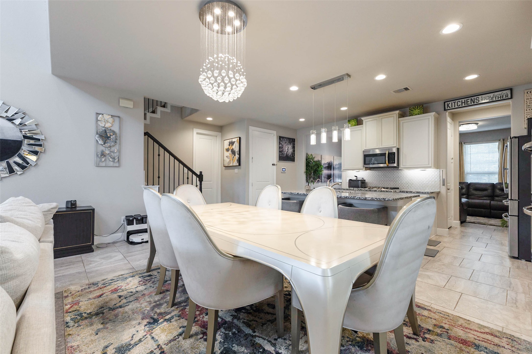The dining area sits beside the kitchen and under a sparkling chandelier, with enough room for a large table and chairs for the whole family. A staircase with dark iron balusters rises just behind, and the island and bar stools are visible on the right. Recessed lighting keeps the area bright for meals and homework alike. Having the table this close to the kitchen makes serving and cleanup easy.
