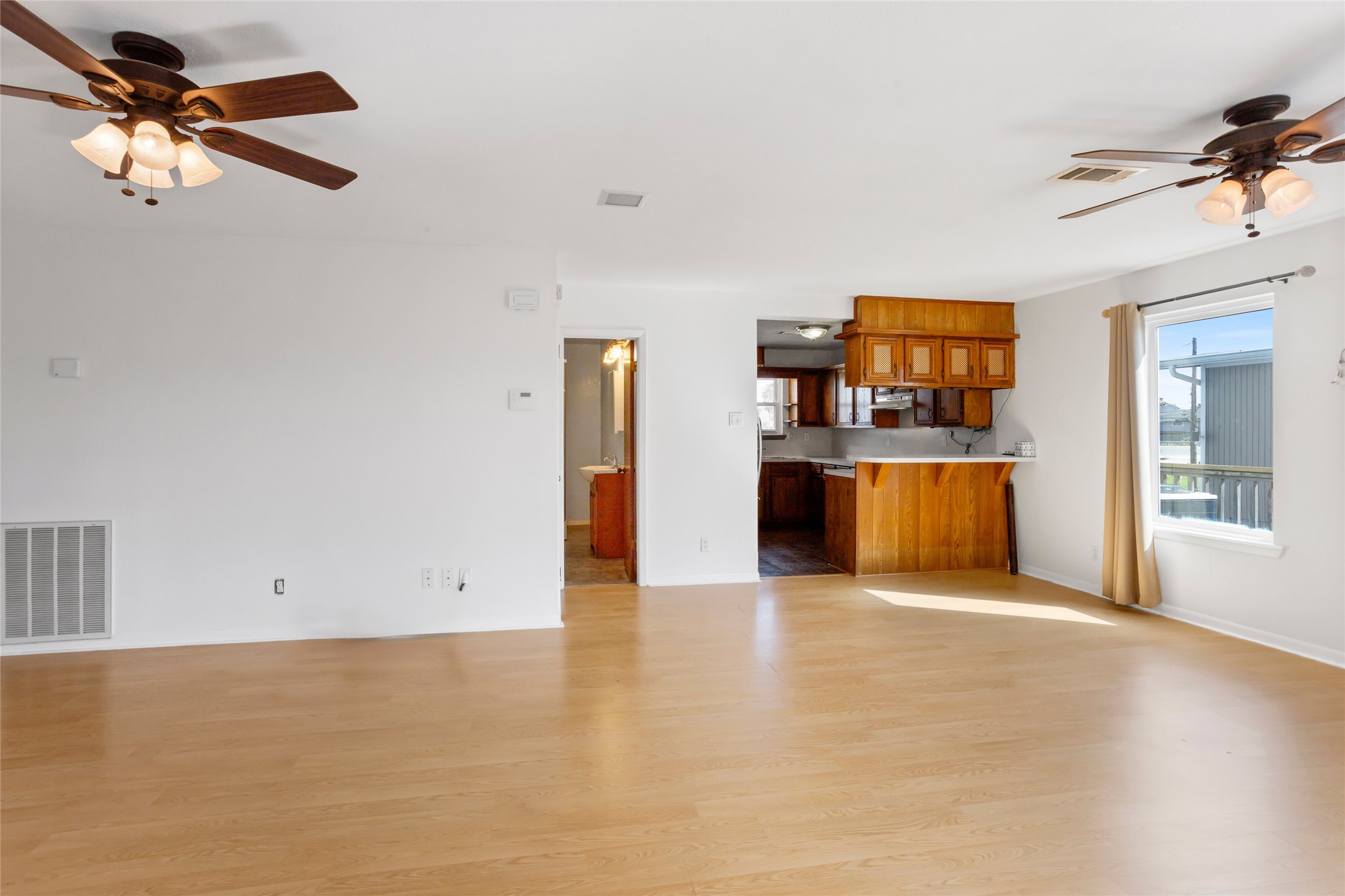 This spacious, light-filled living area features wood laminate flooring, ceiling fans, and a large window. It seamlessly connects to an open kitchen with wooden cabinetry and a breakfast bar, creating a warm and inviting atmosphere. This spacious, light-filled living area features wood laminate flooring, ceiling fans, and a large window. It seamlessly connects to an open kitchen with wooden cabinetry and a breakfast bar, creating a warm and inviting atmosphere.