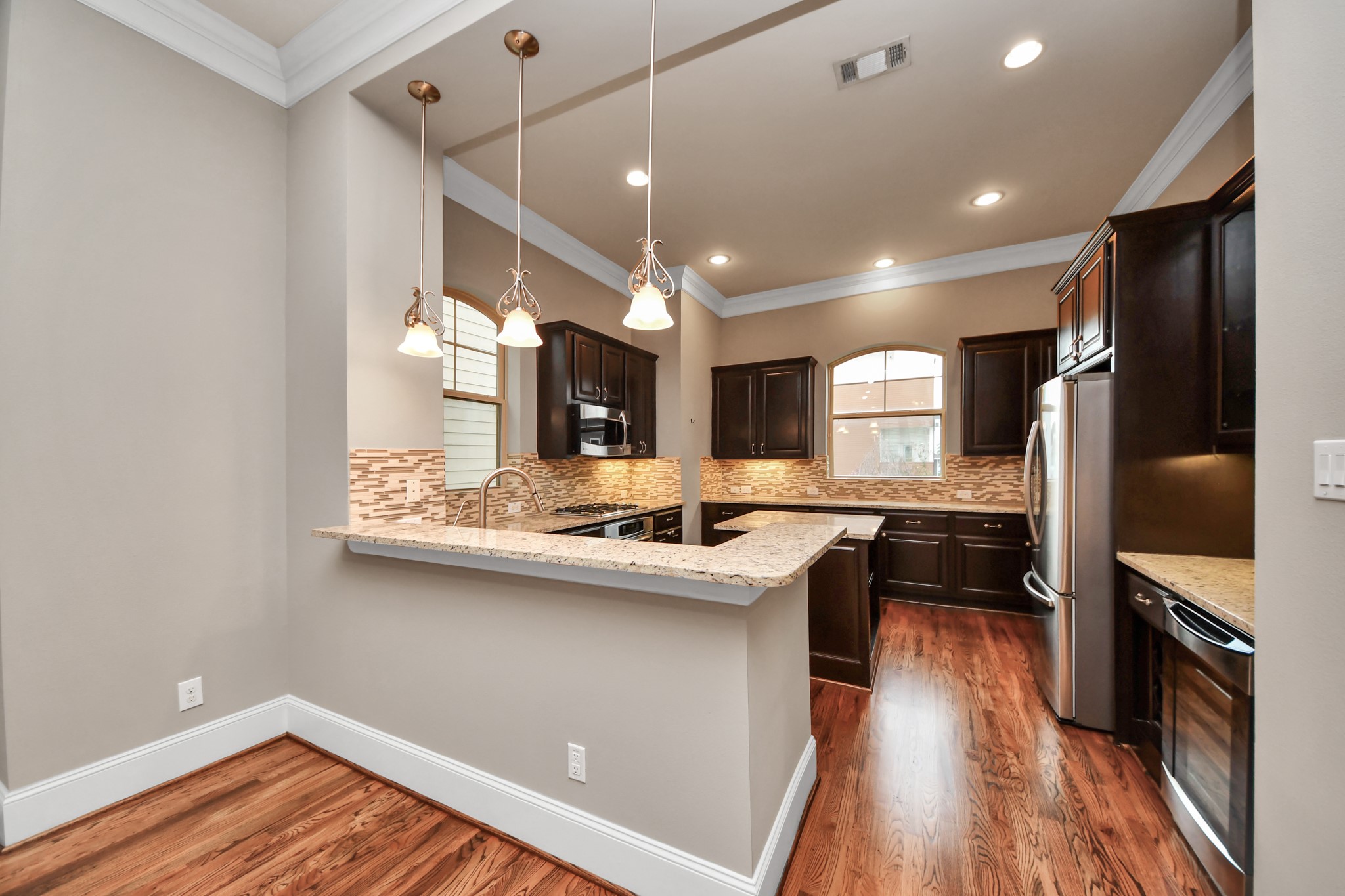 Elegant kitchen featuring rich wood cabinetry, granite countertops, and modern stainless steel appliances. The open layout includes a breakfast bar with pendant lighting, complemented by warm hardwood floors and ample natural light from arched windows.