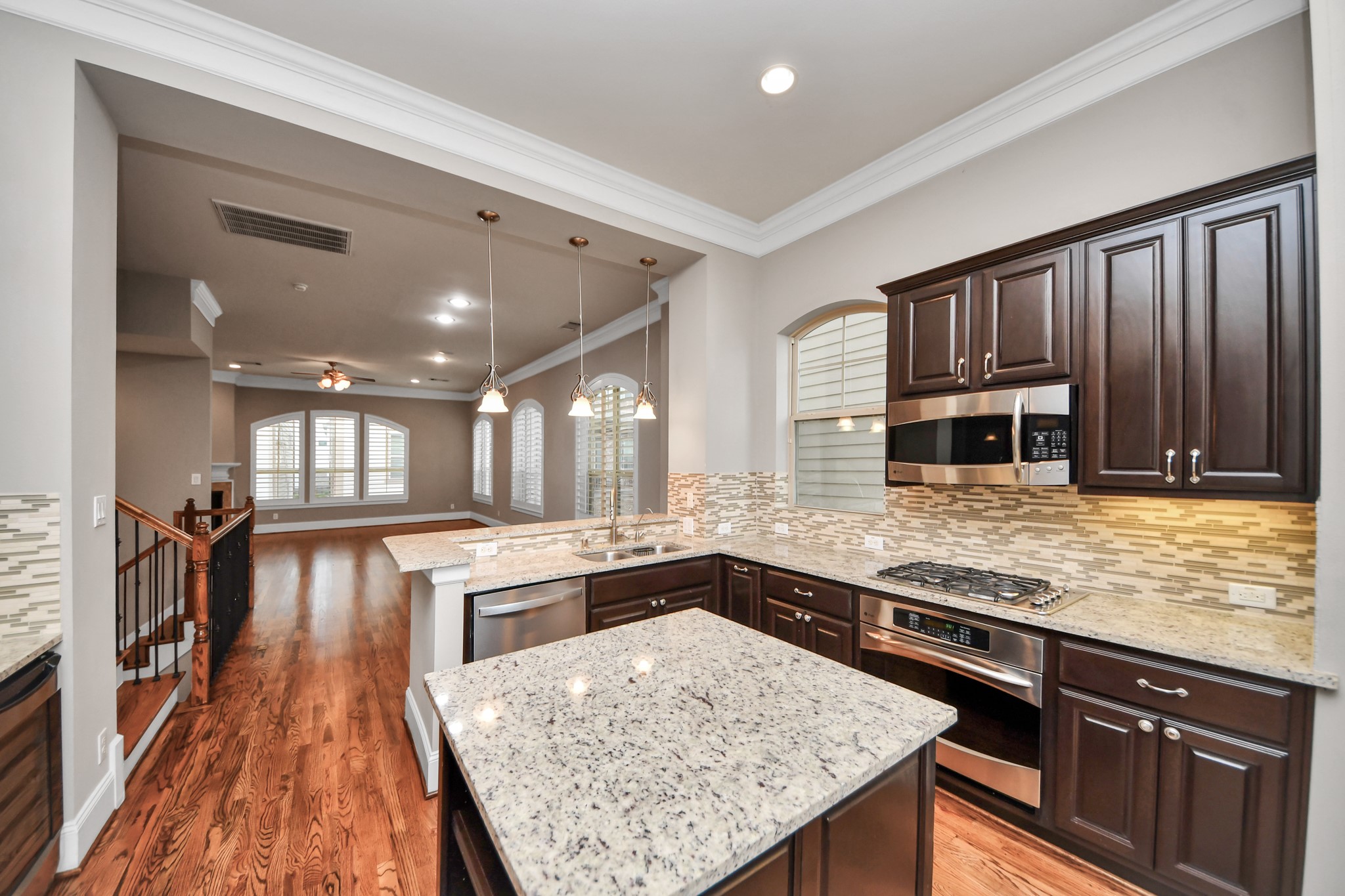 Elegant open-concept kitchen with rich wood cabinetry, granite countertops, and stainless steel appliances, seamlessly flowing into a spacious living area with hardwood floors and large arched windows.