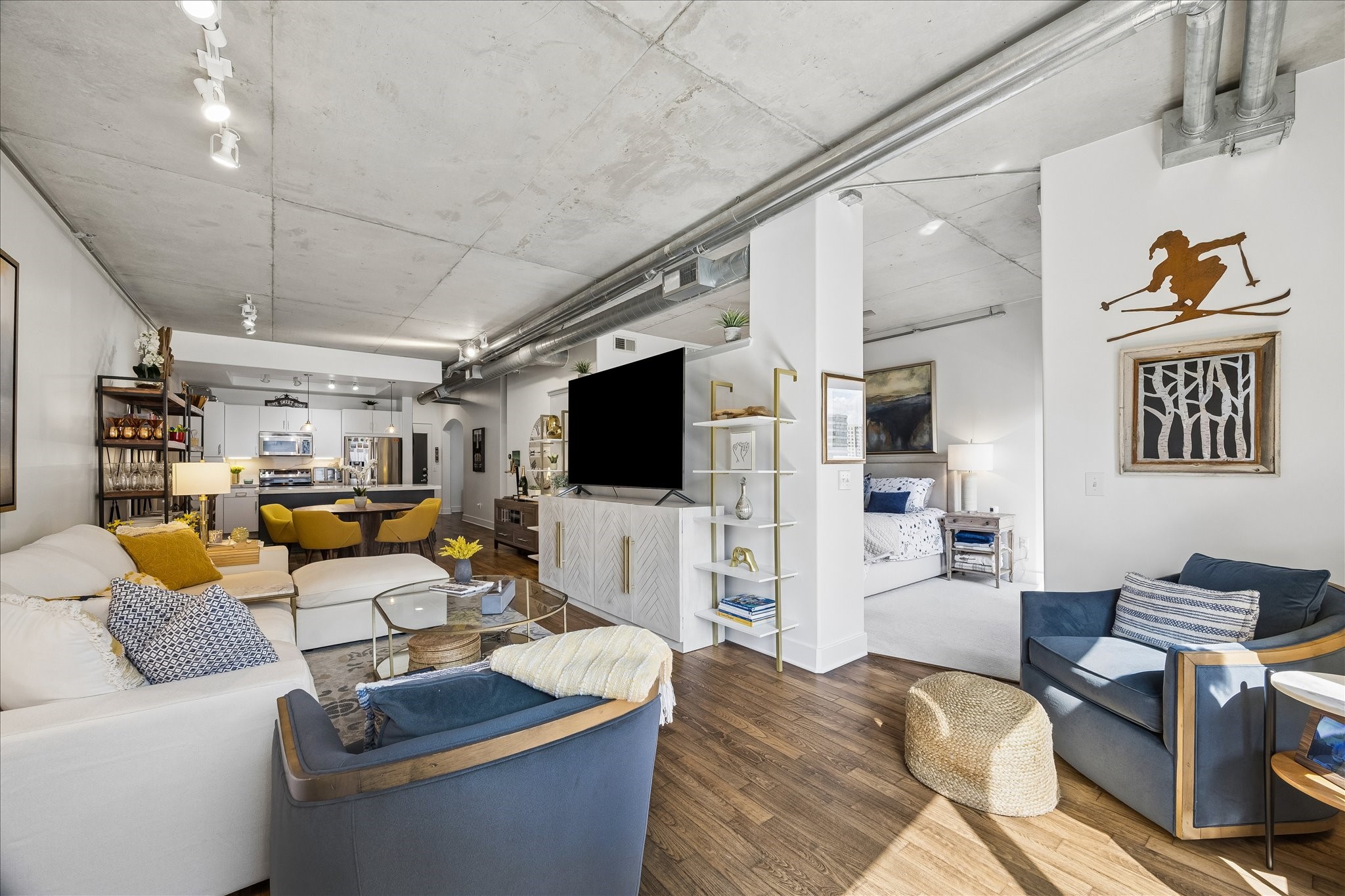 Living area with exposed concrete ceilings, sleek hardwood floors, and abundant natural light. The space seamlessly flows into a contemporary kitchen and cozy bedroom nook, creating an urban retreat.