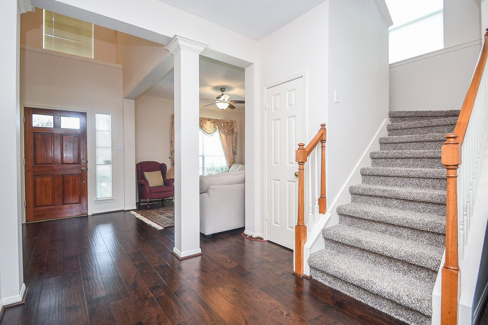 This entryway features beautiful dark hardwood floors, a wooden front door with sidelights, and a carpeted staircase. The space opens into a bright living area with a ceiling fan, offering a warm and inviting atmosphere.