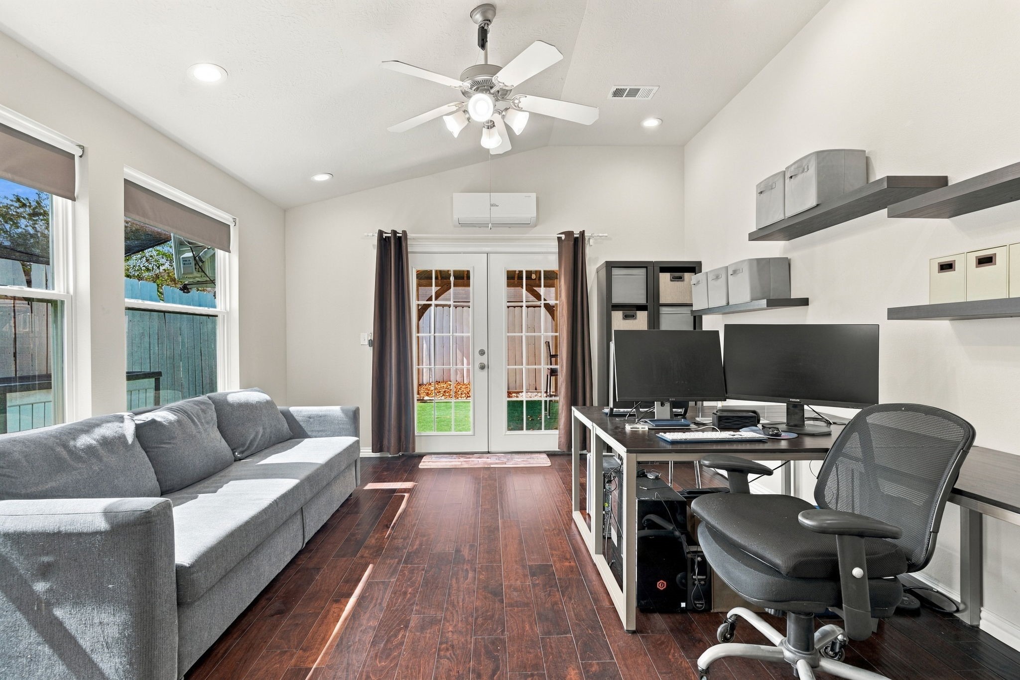 Bright and spacious office area featuring elegant wood flooring, a comfortable gray sofa, and ample natural light streaming through large windows and glass doors.