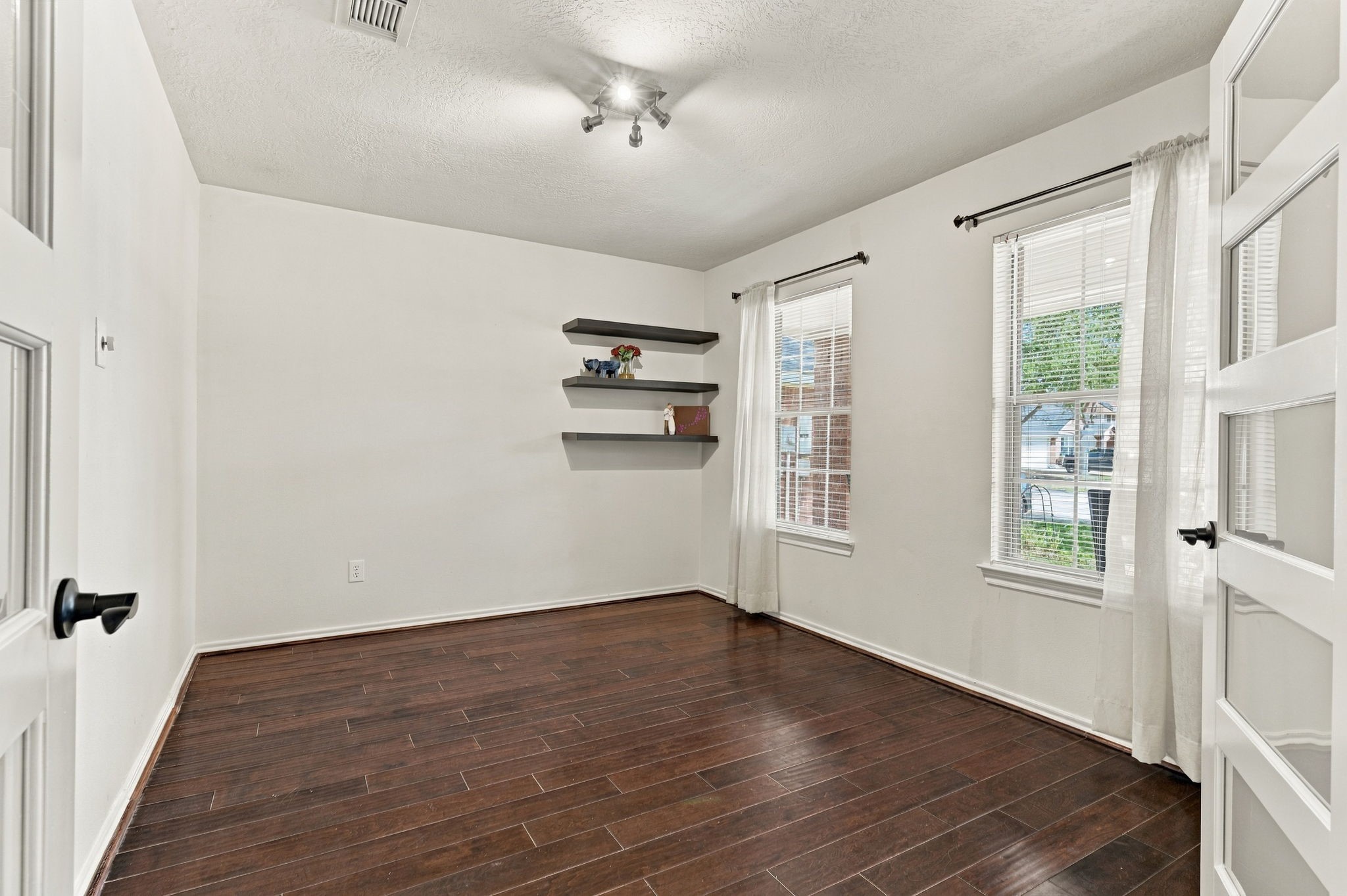 Bright and inviting room featuring rich hardwood floors, ample natural light from large windows, and stylish floating shelves for a modern touch.